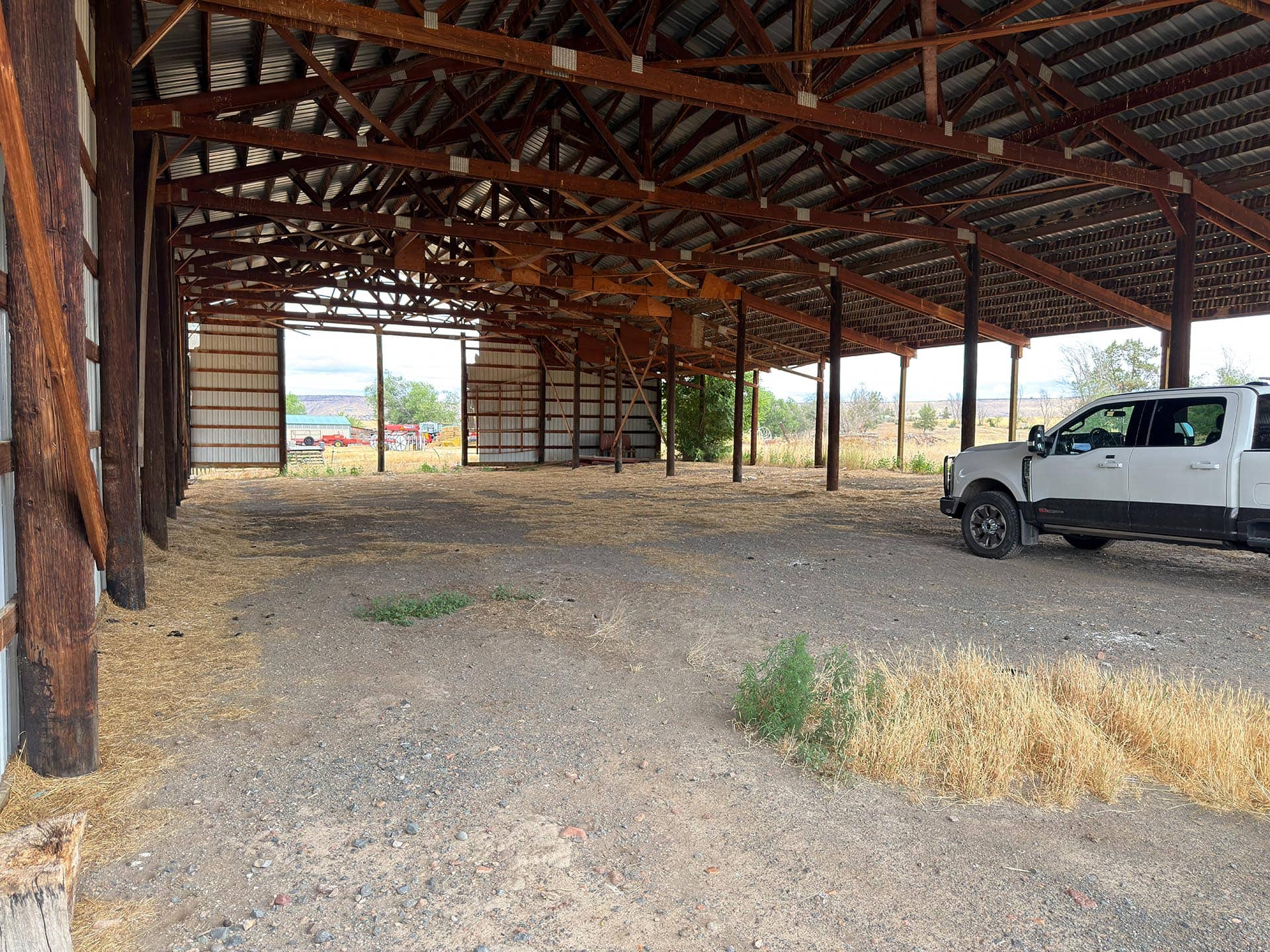 Open-sided barn interior with exposed wooden trusses and a white pickup truck parked on dirt ground to the right.