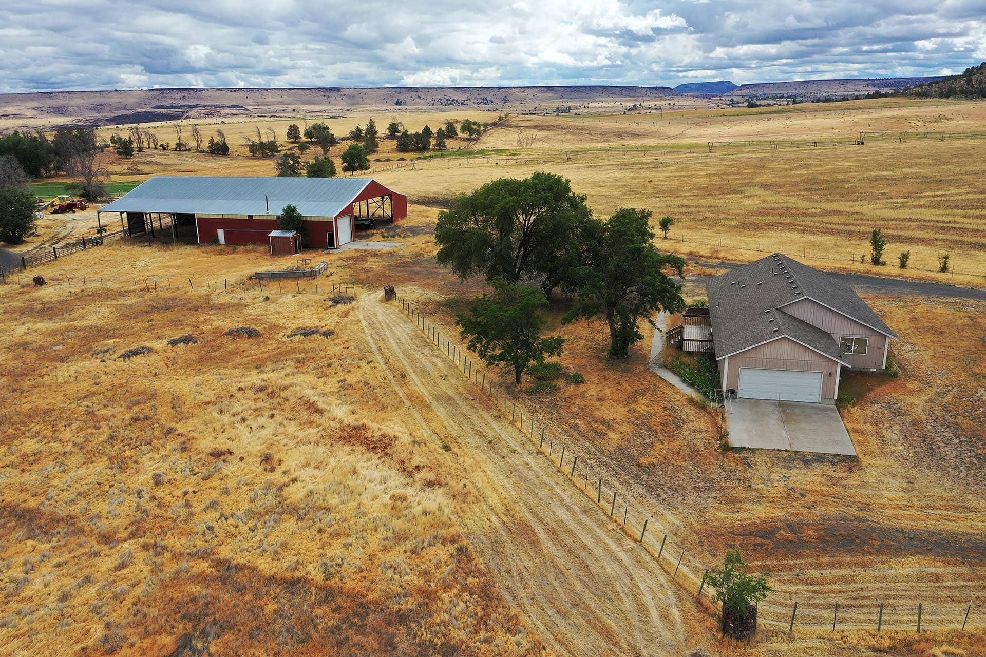 Aerial view of a farm: red barn with a metal roof beside a gray-roofed house, set in dry golden grassland with dirt roads and fences.