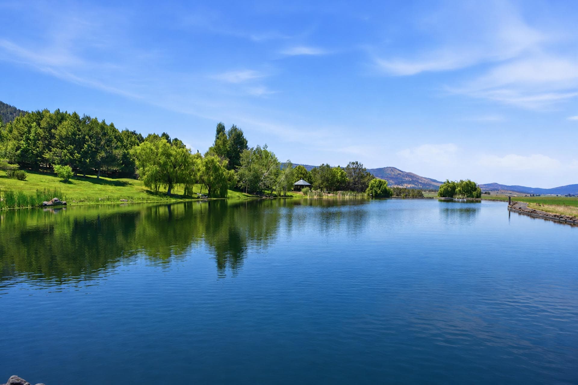 Serene lake bordered by green trees, reflecting a clear blue sky and distant hills.