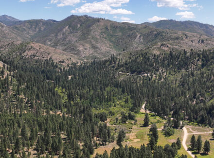 Panoramic view of rugged, forested hills and distant mountains under a blue sky with scattered clouds, a winding dirt road in the valley.