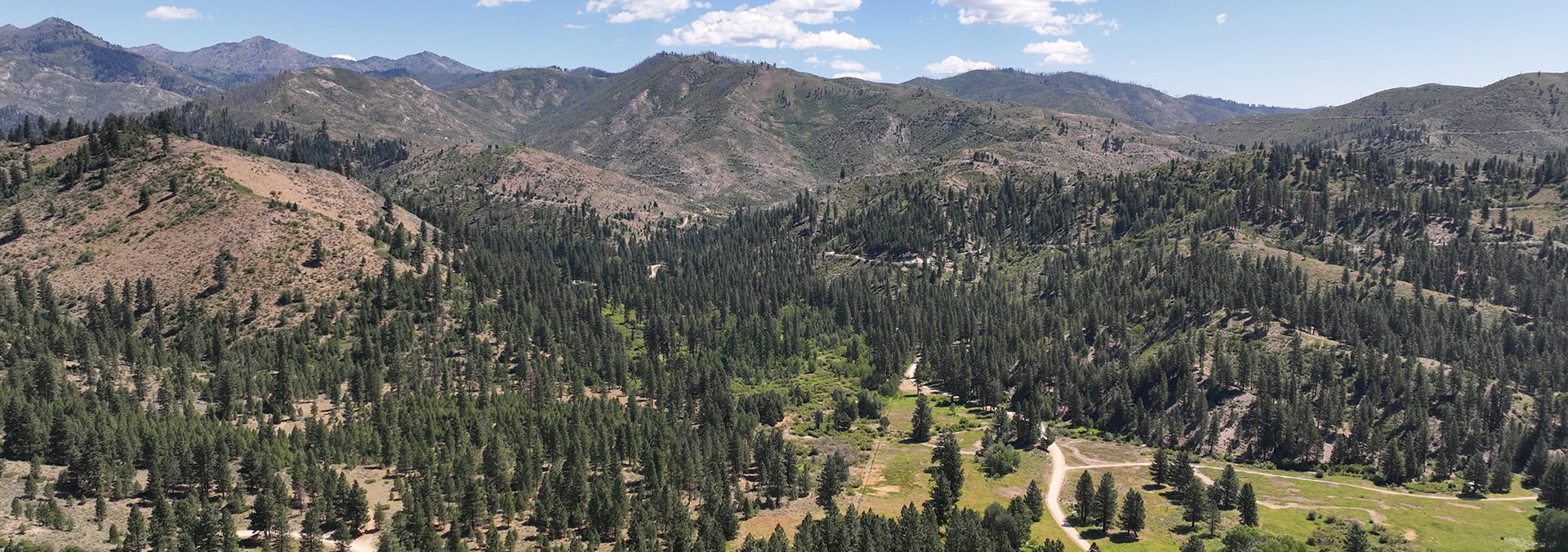 Panoramic view of rugged, forested hills and distant mountains under a blue sky with scattered clouds, a winding dirt road in the valley.