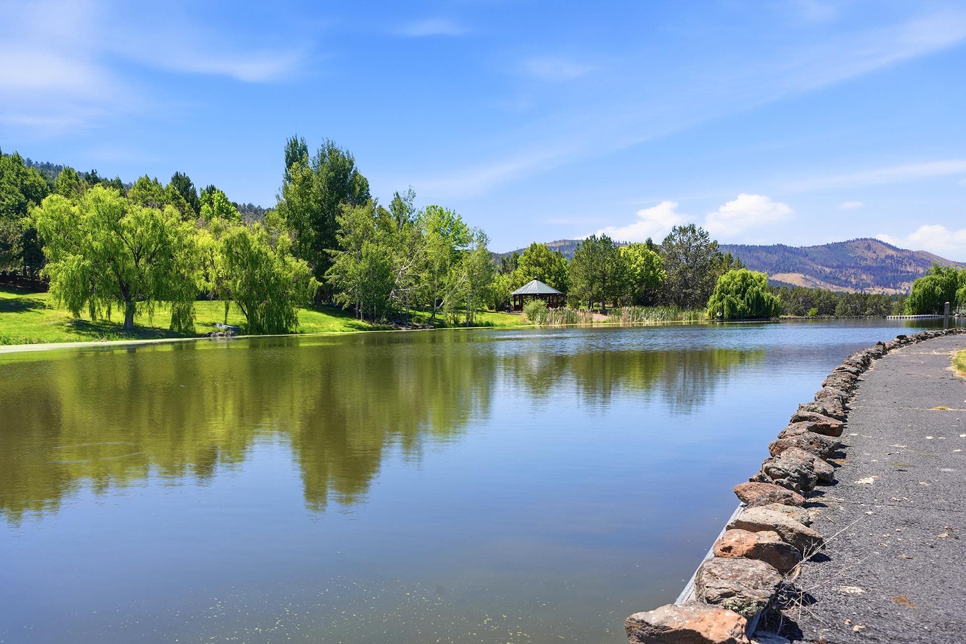 Calm lake reflecting green trees along a grassy bank, with a gazebo and distant mountains under a clear blue sky.