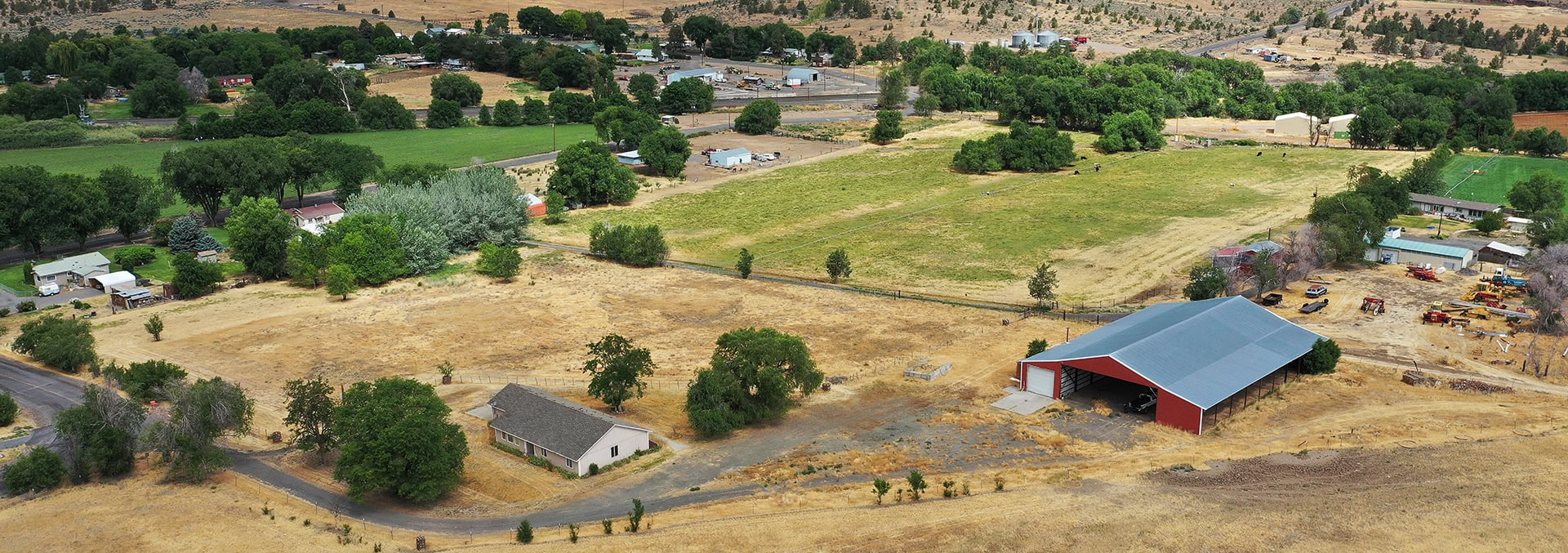 Aerial view of a rural farm: red barn with a large metal roof, a smaller shed, dry golden fields, and scattered houses and trees nearby.