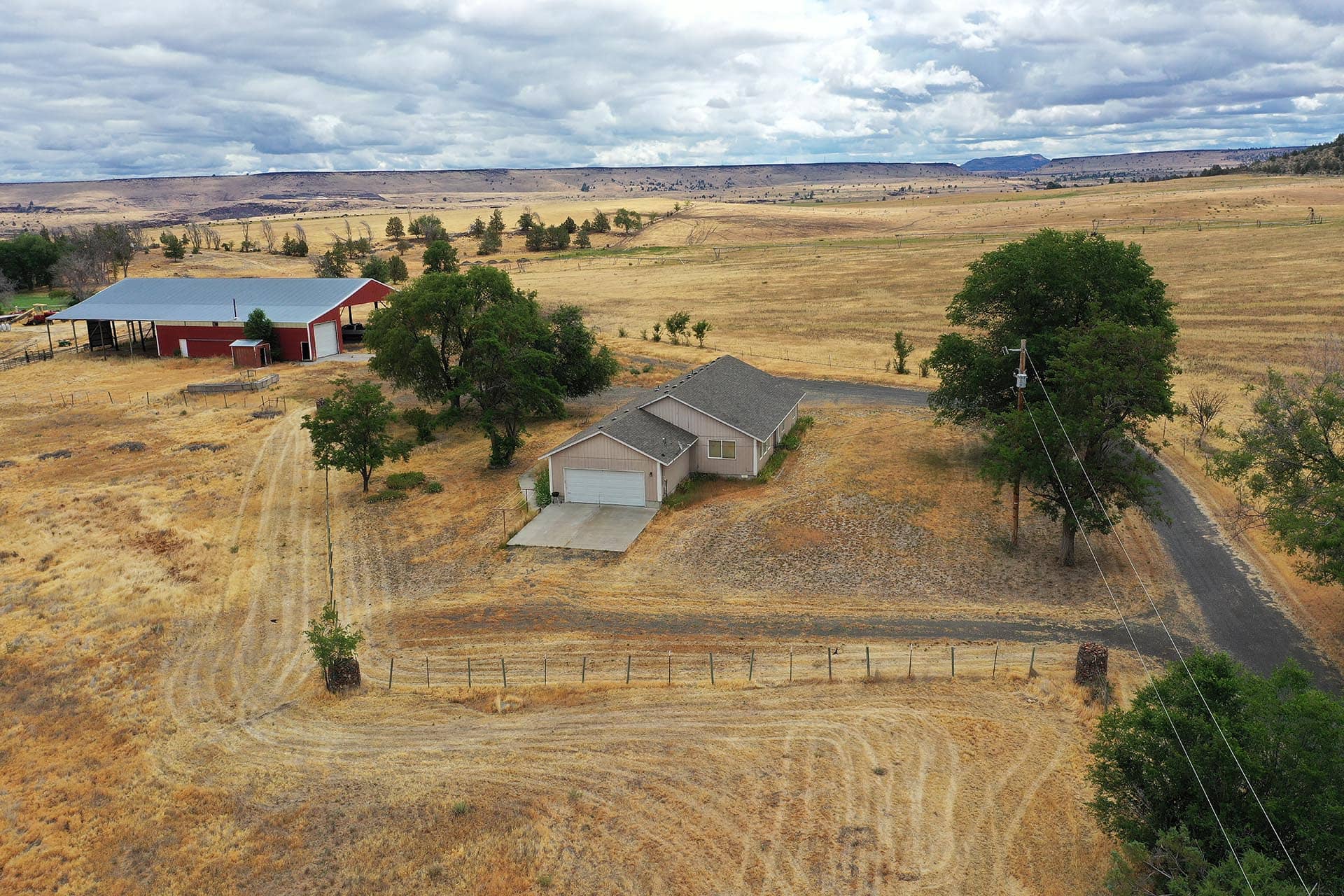 Aerial view of a rural property with a beige house, red barn, dry brown fields, and scattered trees under a cloudy sky.