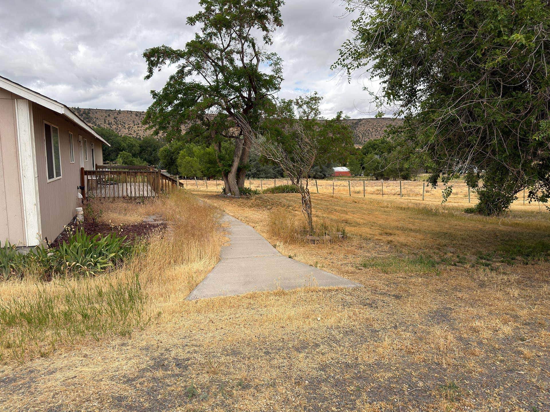 Side of a beige house with a wooden deck on the left, a concrete path, and dry yellow grass in a rural yard under cloudy skies.