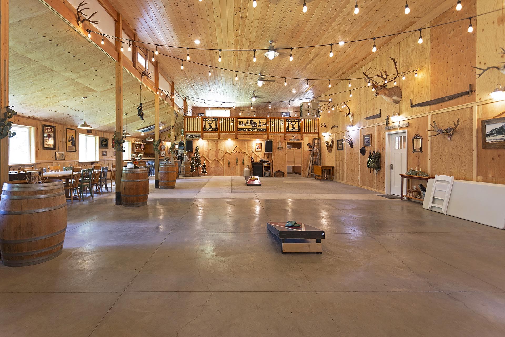 Rustic indoor hall with wood-paneled walls and ceiling, string lights, deer mounts, and wooden barrels near tables along the left side. A loft overlooks the main space.