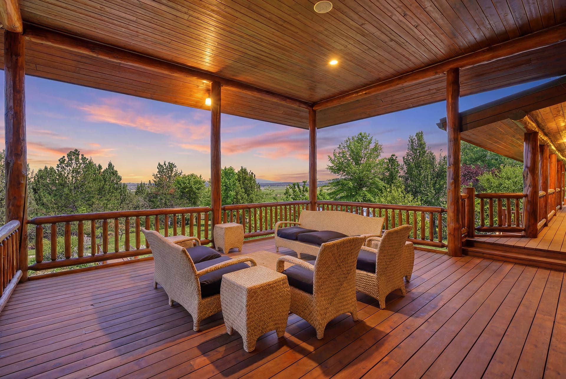 Wooden deck with wicker chairs and a sofa, overlooking trees at sunset.