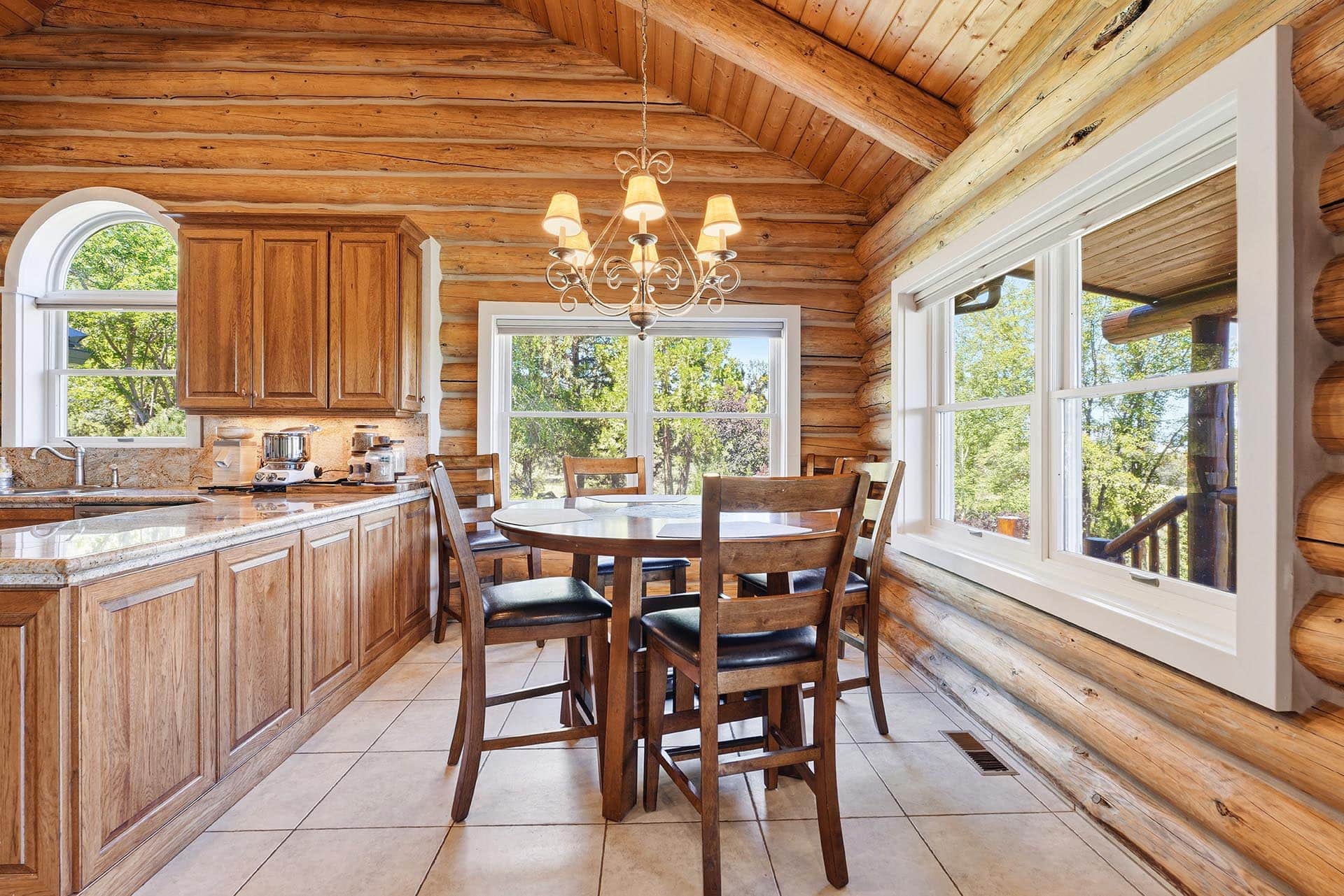 Rustic log cabin kitchen with wooden cabinets, a chandelier, a round dining table, and large windows showing greenery outside.