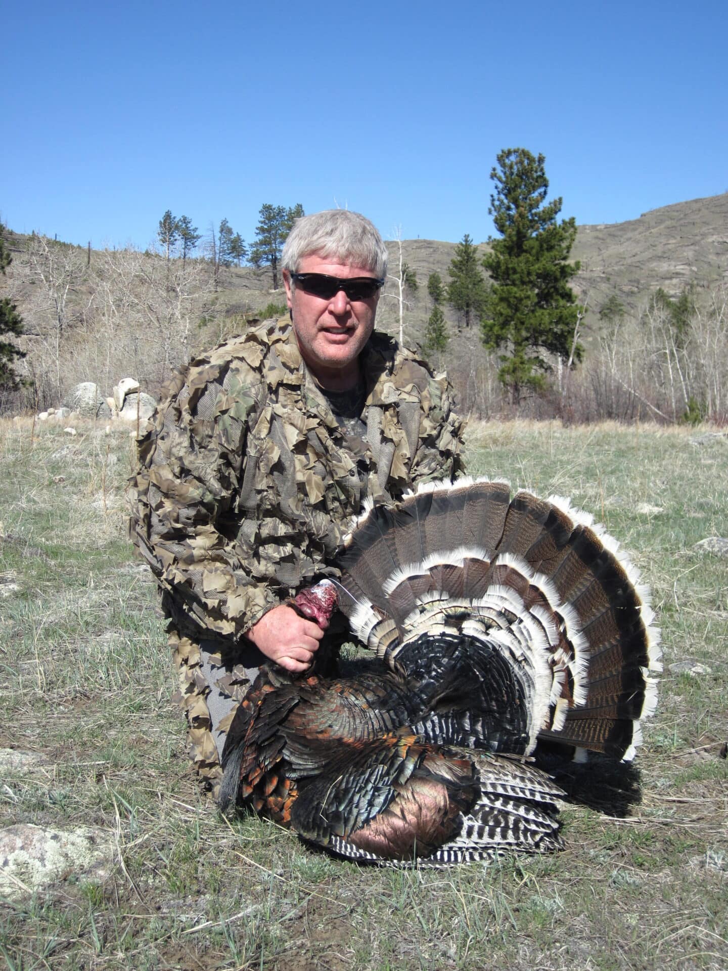 Hunter in camo clothing posing outdoors with a large wild turkey in a grassy field and clear blue sky behind him.