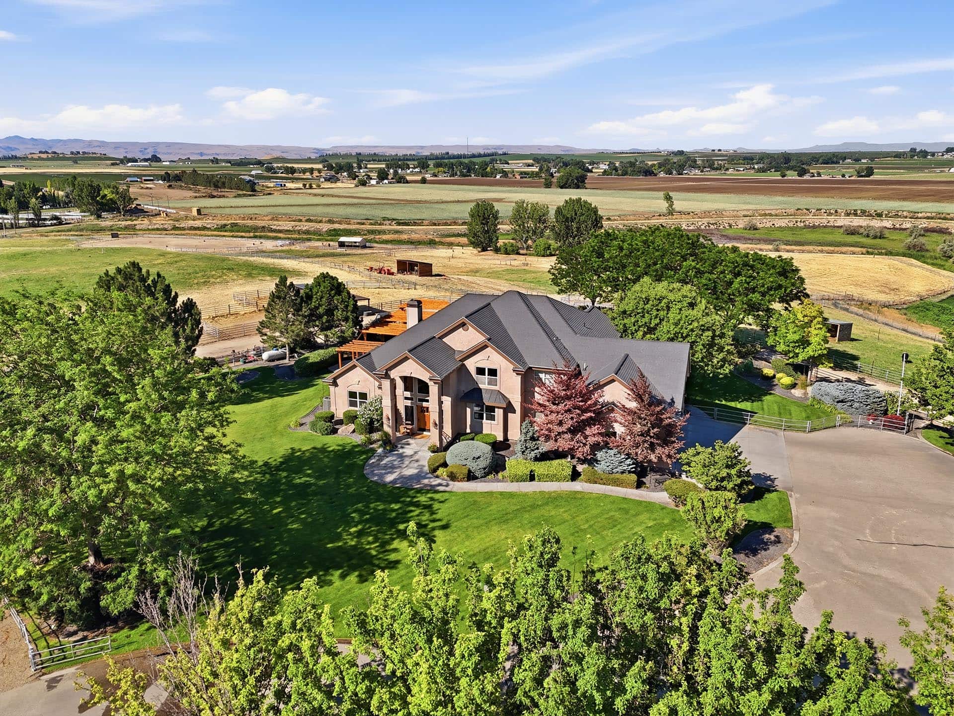 Aerial view of a large brick house with a gray roof, lush green lawn, and mature trees in a rural farmland setting