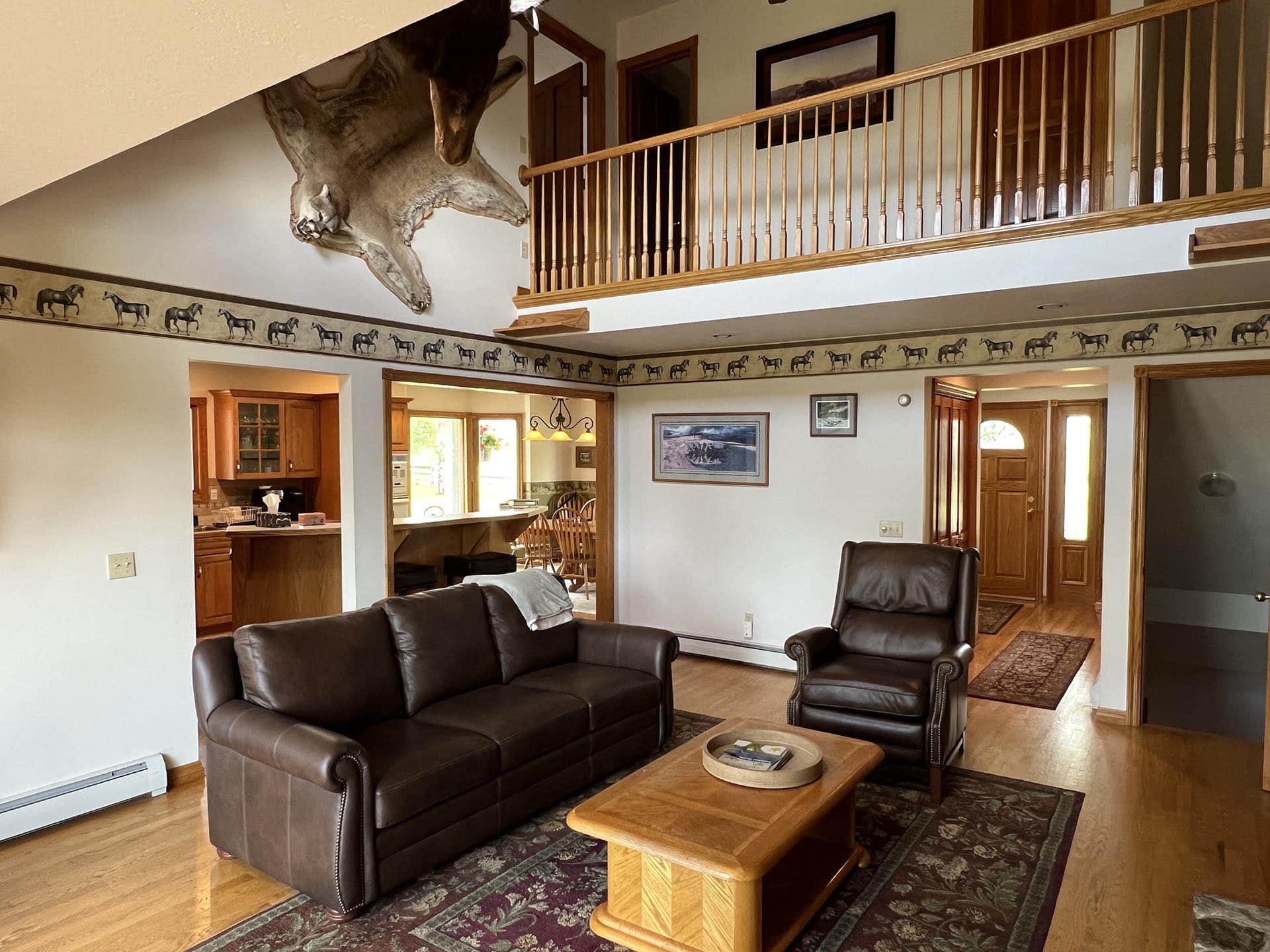 Living room with dark leather sofa set, a wooden coffee table, and a mounted animal head high on the left wall; open kitchen visible in the background and a wooden railing on a second-floor balcony above.