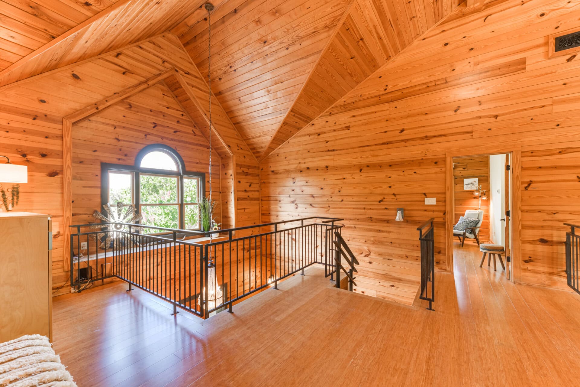 Wood-paneled loft with vaulted ceiling, black metal railing, and an arched window letting in bright light.