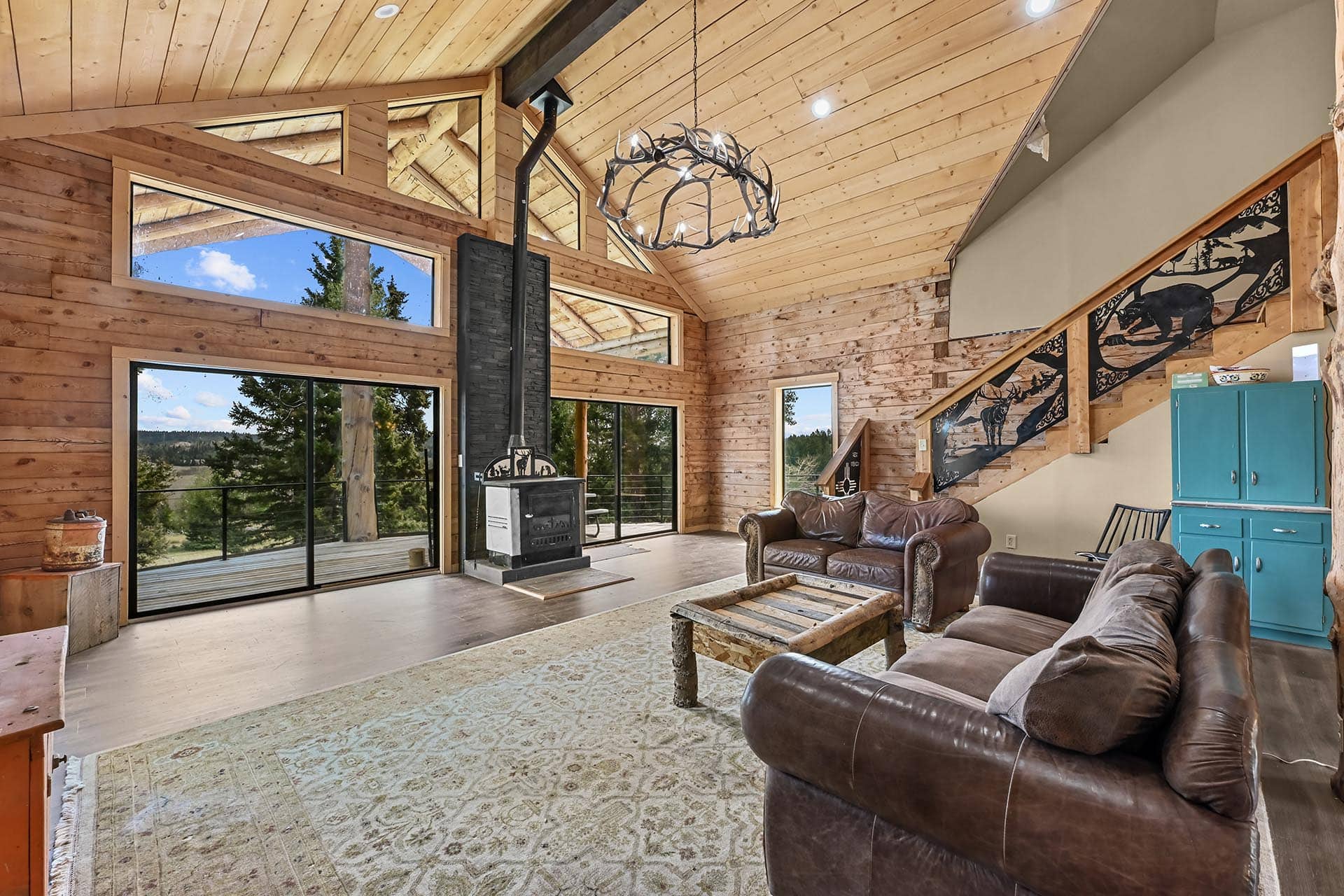 Open living room in a log cabin with large windows, a black wood stove, antler chandelier, and brown leather sofas surrounding a rustic coffee table.