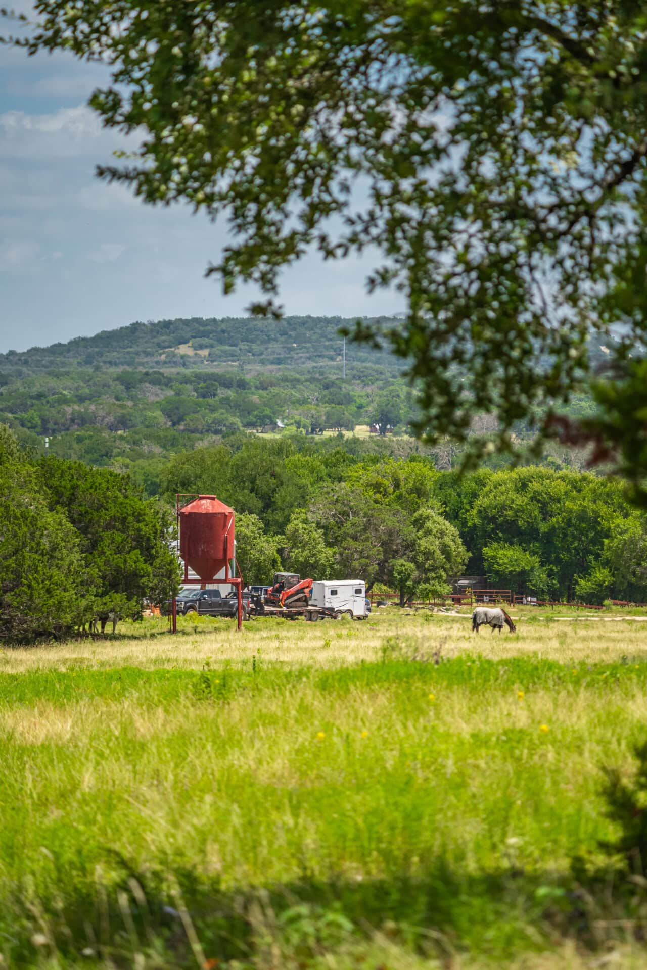 Wide rural scene: grassy field in the foreground with a red grain silo and farm equipment near a line of trees; rolling hills in the distance under a blue sky.