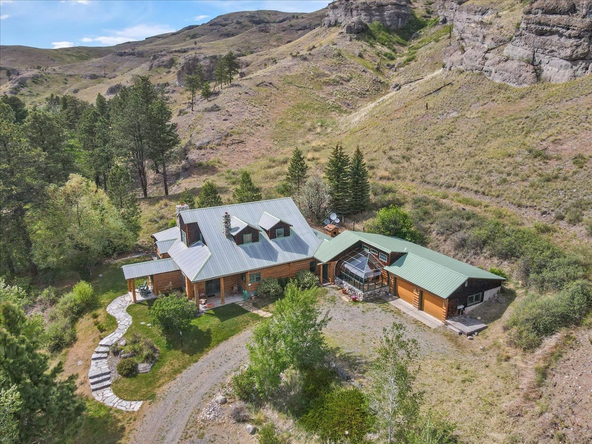 Aerial view of a multi-building wooden cabin complex on a grassy hillside with trees and rocky cliffs in the background, connected by stone paths and a greenhouse.