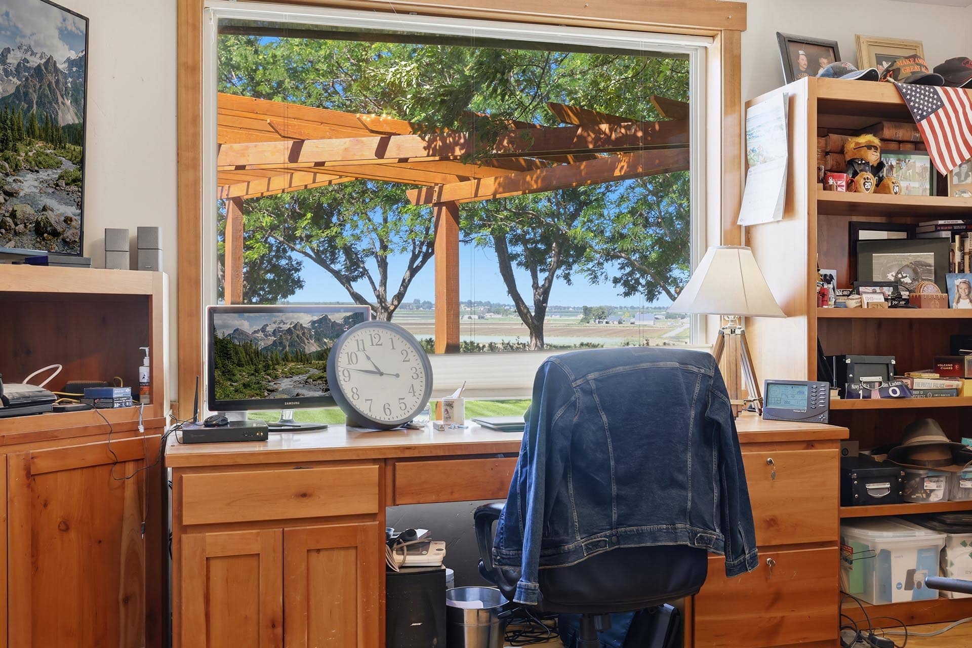 Home office scene with a large window view of trees and wooden pergola, a desk clock, a computer monitor showing mountains, and a denim-covered chair back.