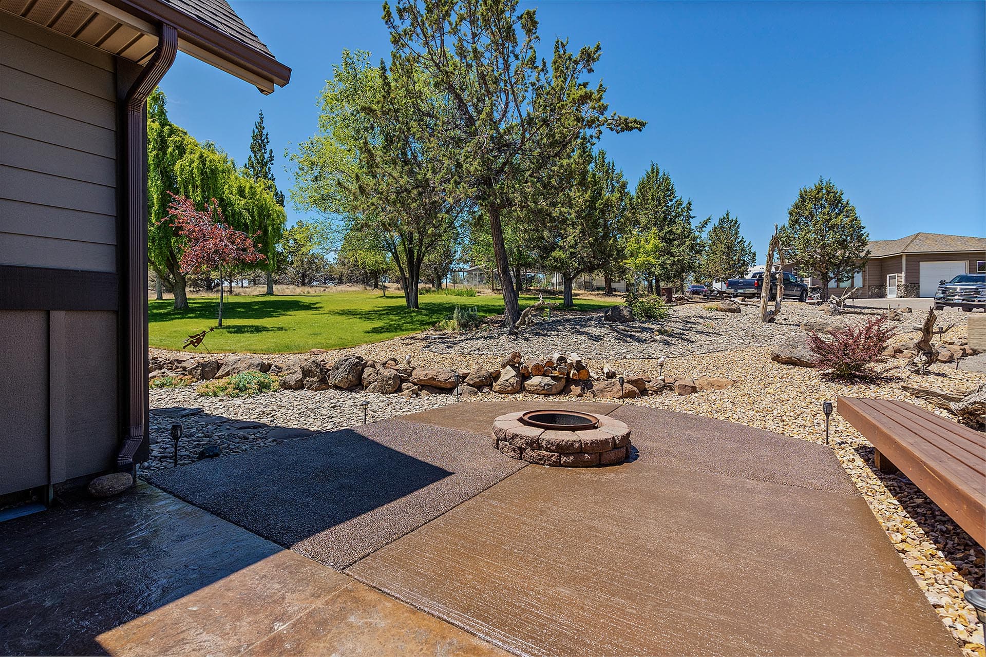Backyard with a circular stone fire pit on a paved patio, gravel landscaping, and green trees under a blue sky.