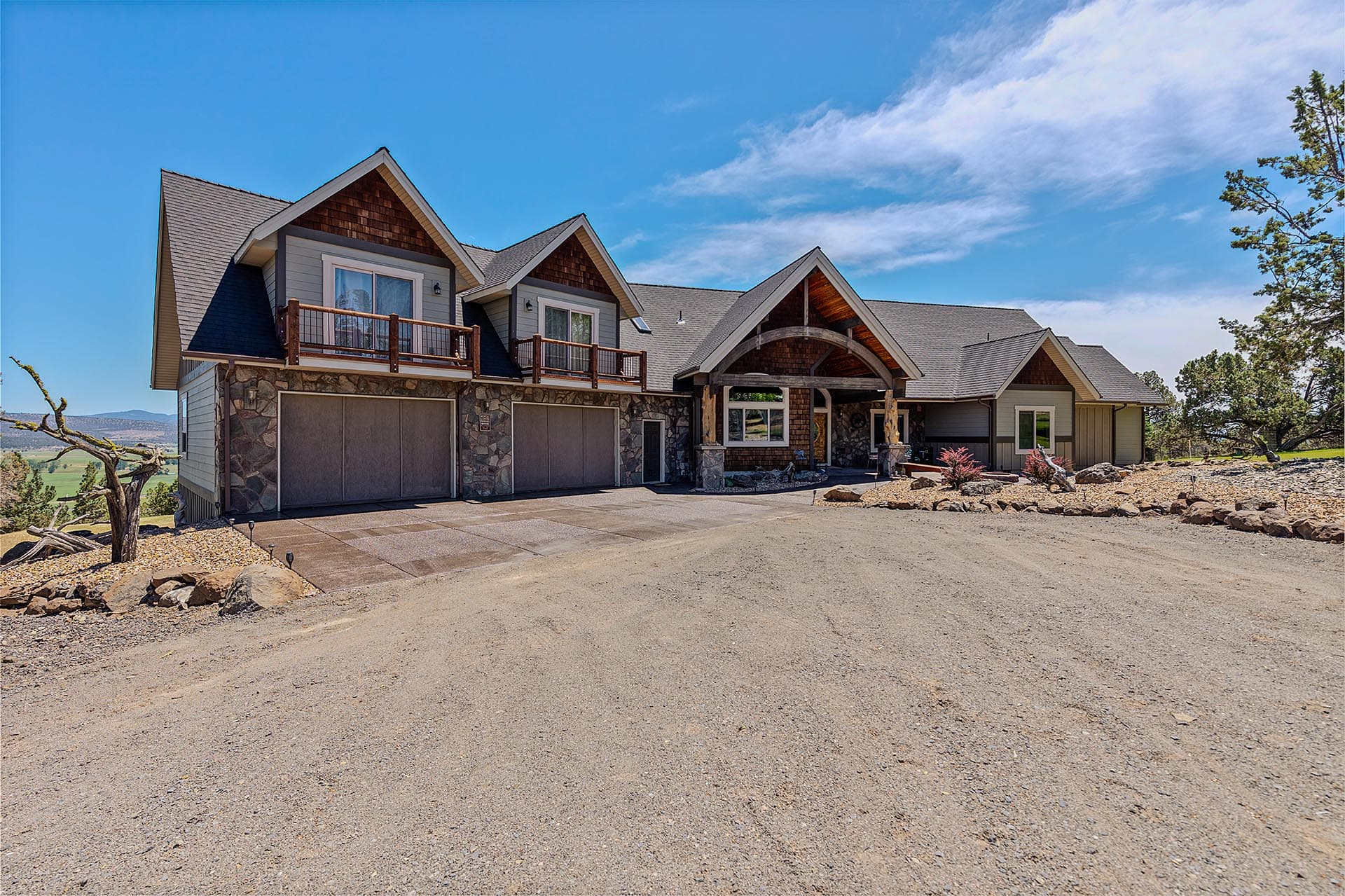 Modern two-story home with stone and wood facade, balconies, and a three-car garage on a gravel driveway beneath a bright blue sky.