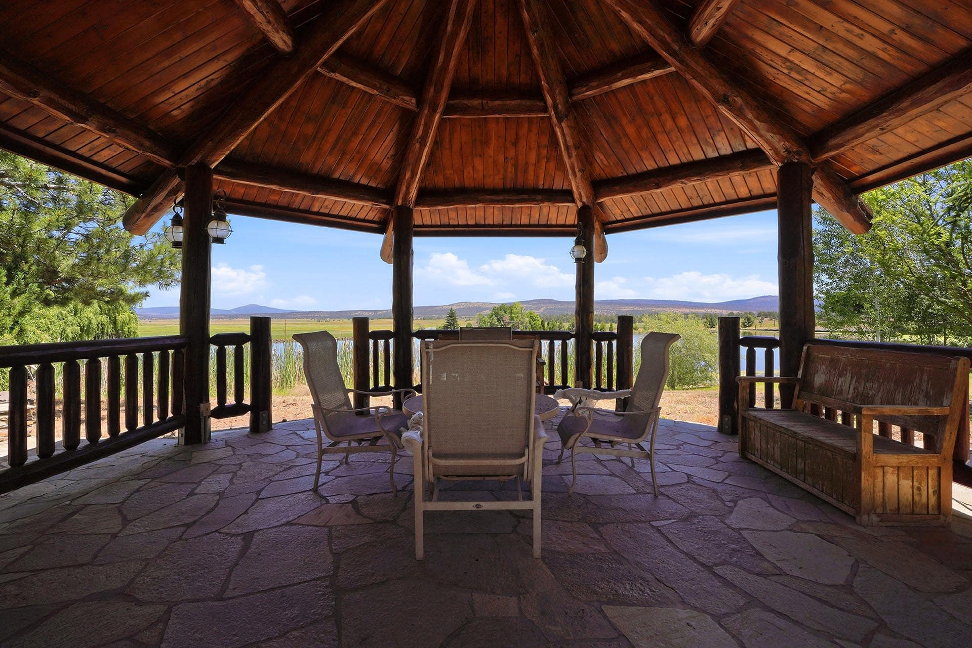 Wooden gazebo with stone floor and six chairs around a table, overlooking a lake and distant hills.