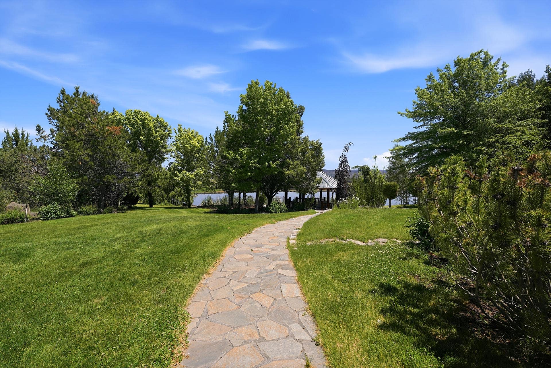 Stone walkway through a green park toward a lakeside gazebo under a bright blue sky.