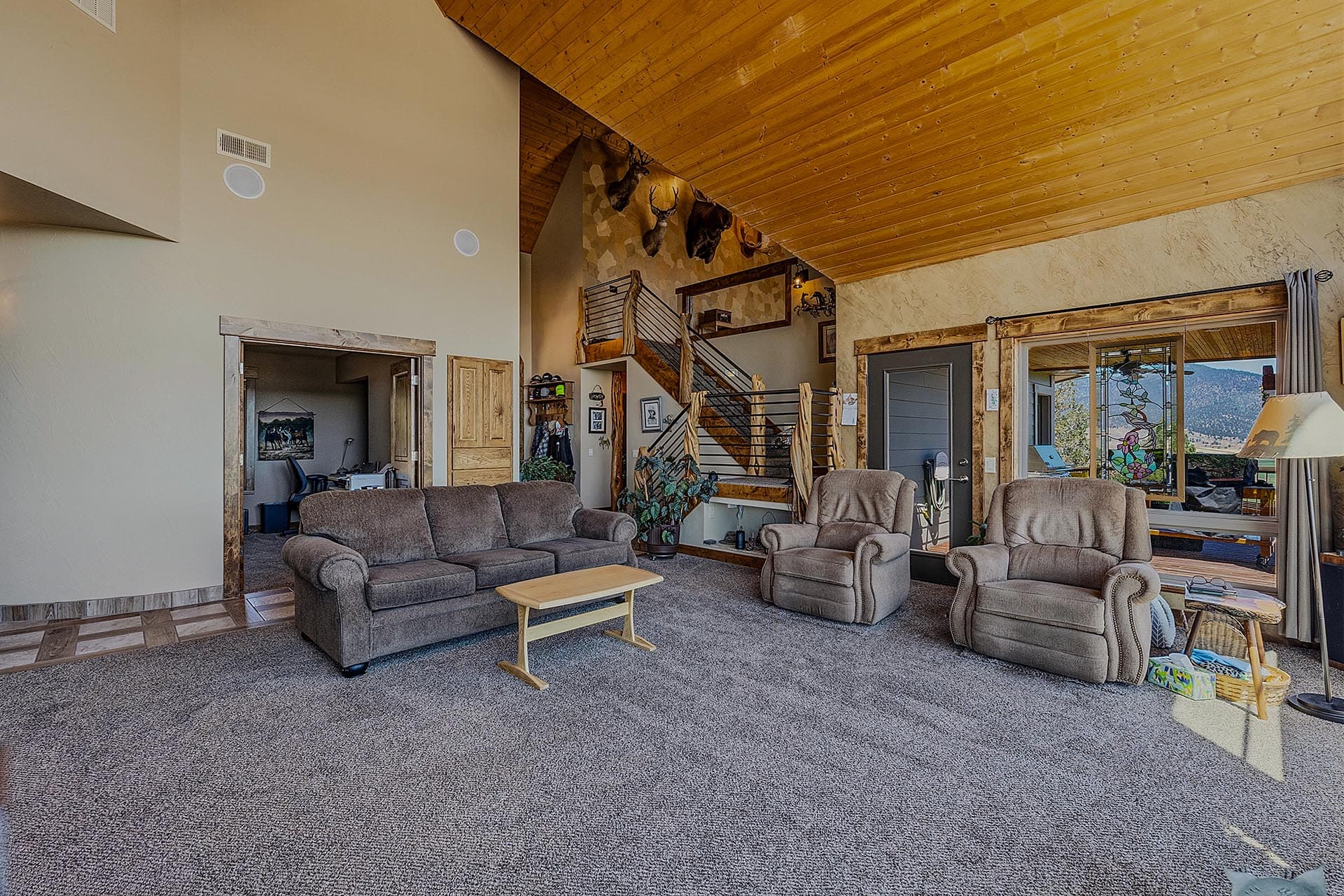 Spacious living room with a high wooden ceiling, stone accent wall, and brown sofas facing a windowed wall showing mountains outside through a stained glass panel.