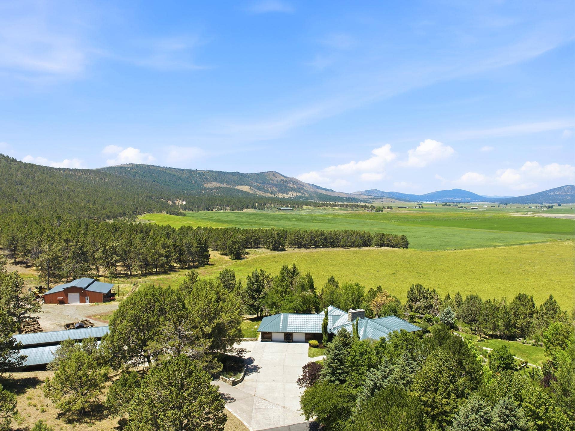 Aerial view of a rural landscape with green fields, pine trees, and distant hills under a bright blue sky.