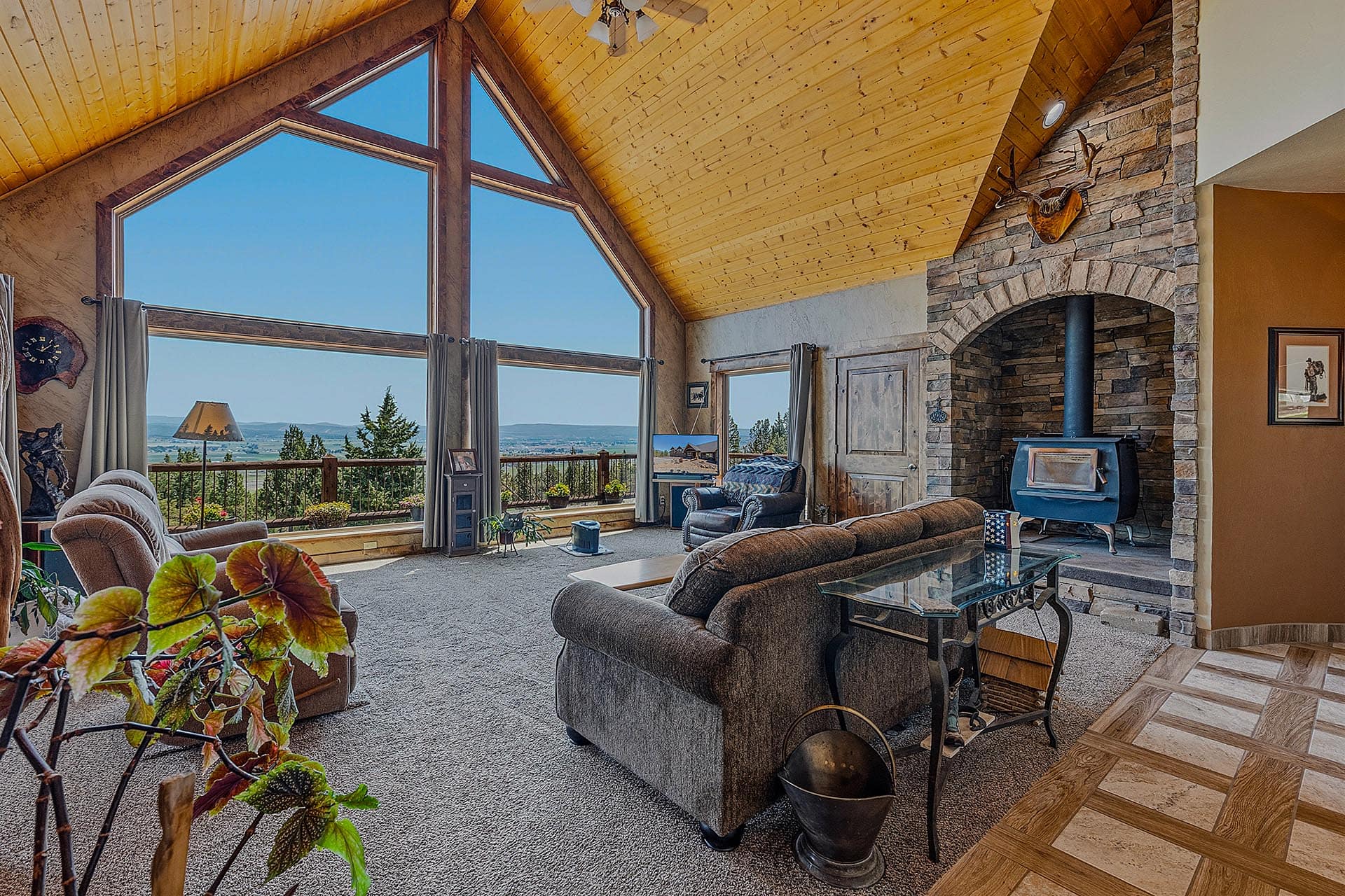 Cozy living room with a stone fireplace and wood stove, vaulted wooden ceiling, and large triagonal windows offering a lakeside view.