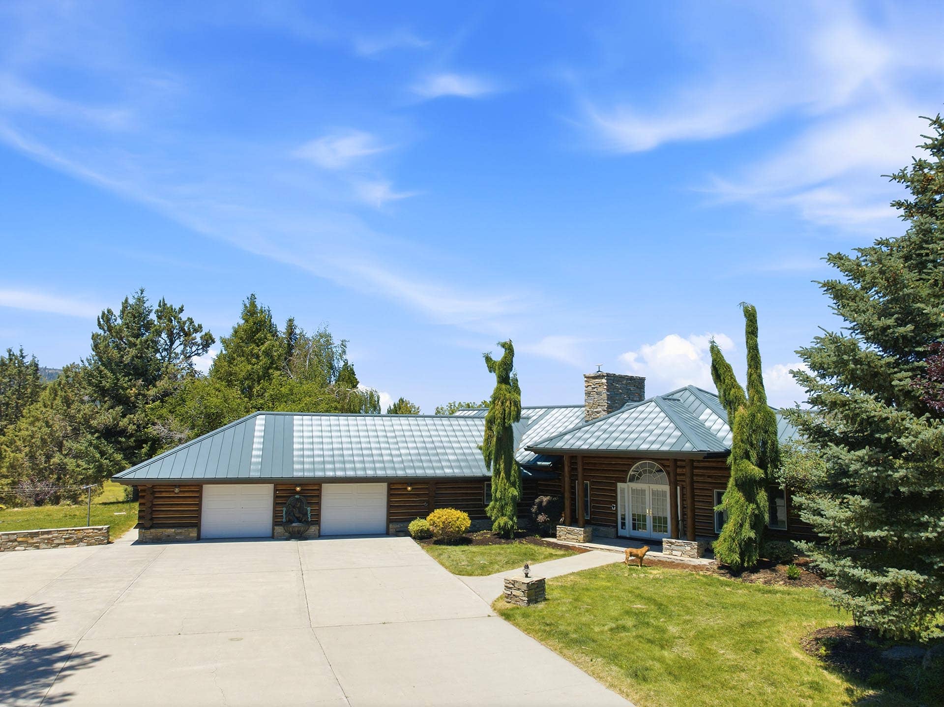 Log cabin-style house with a metallic green roof, three garage doors, and front yard under a blue sky with trees surrounding the property