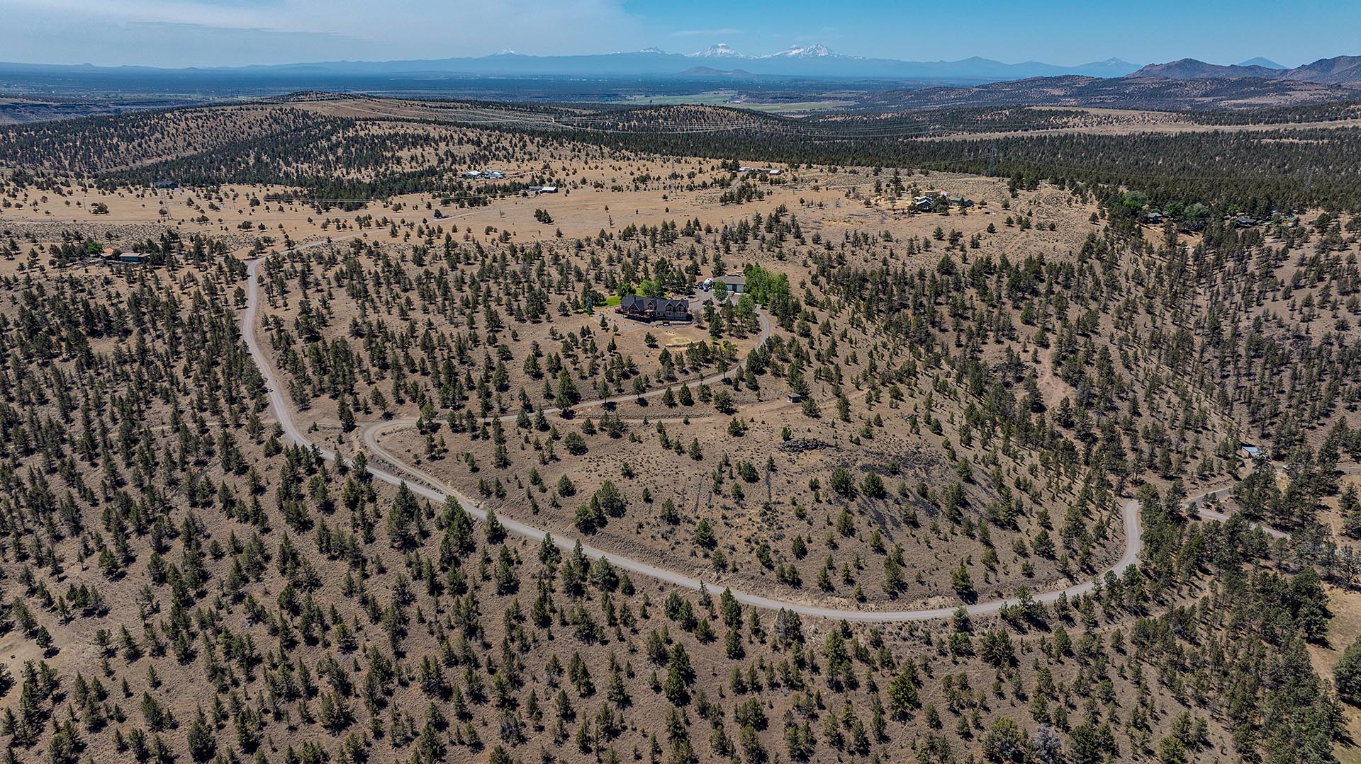 Aerial view of a sparsely wooded, arid landscape with a winding road leading to a large house surrounded by trees at the center and distant mountains on the horizon.