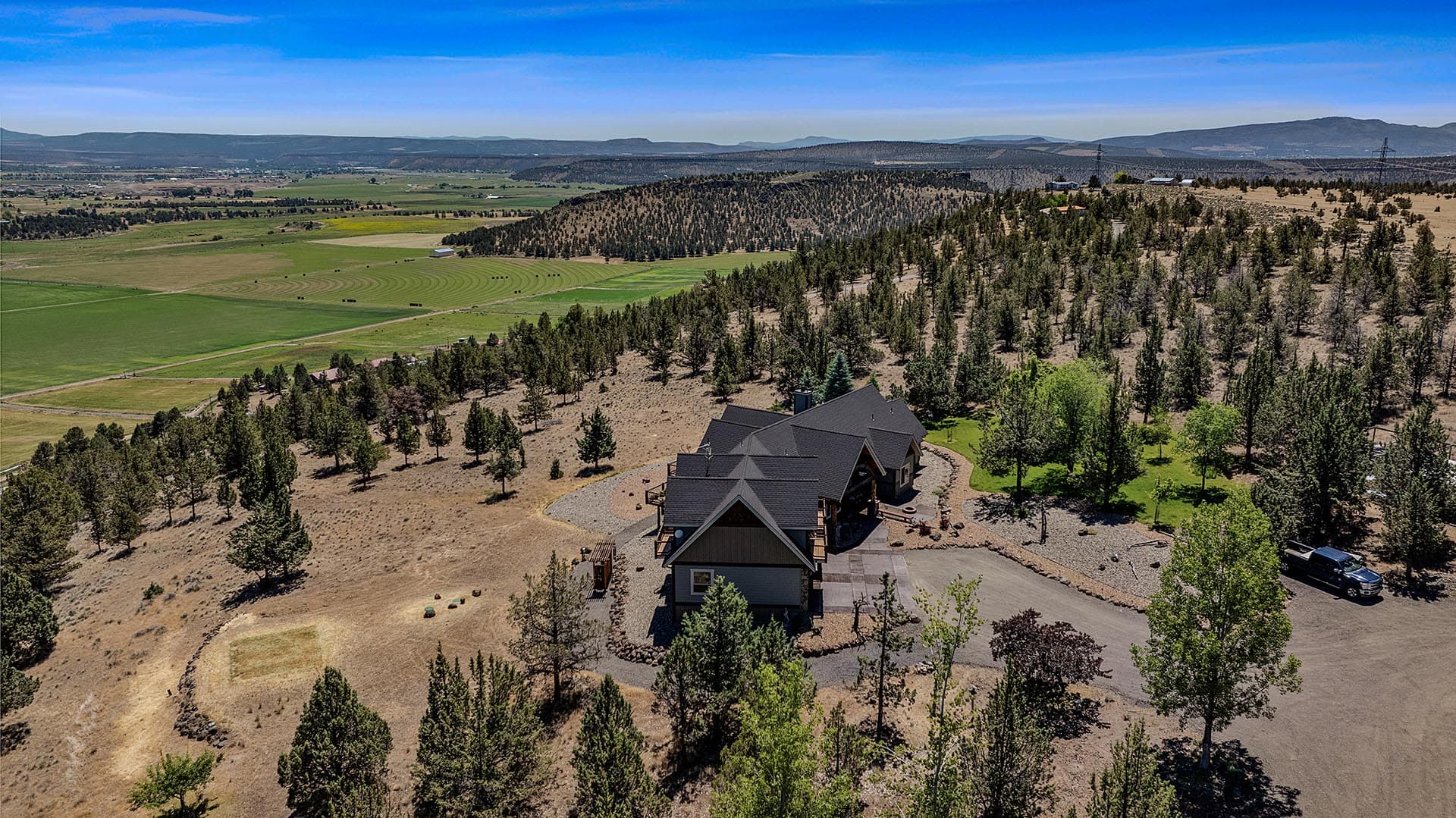 A large house with a dark roof on a forested hillside, with green fields and distant mountains in the background.