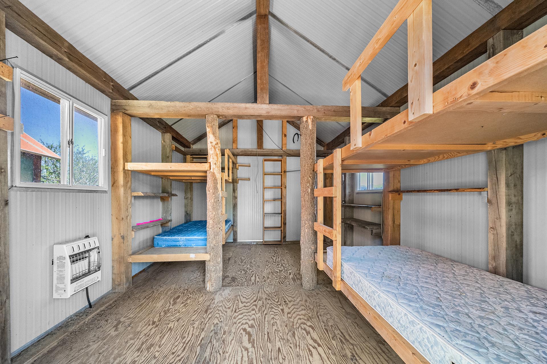 Interior of a rustic bunkhouse with two-tier wooden bunk beds, a window, and a wall heater visible on the left.