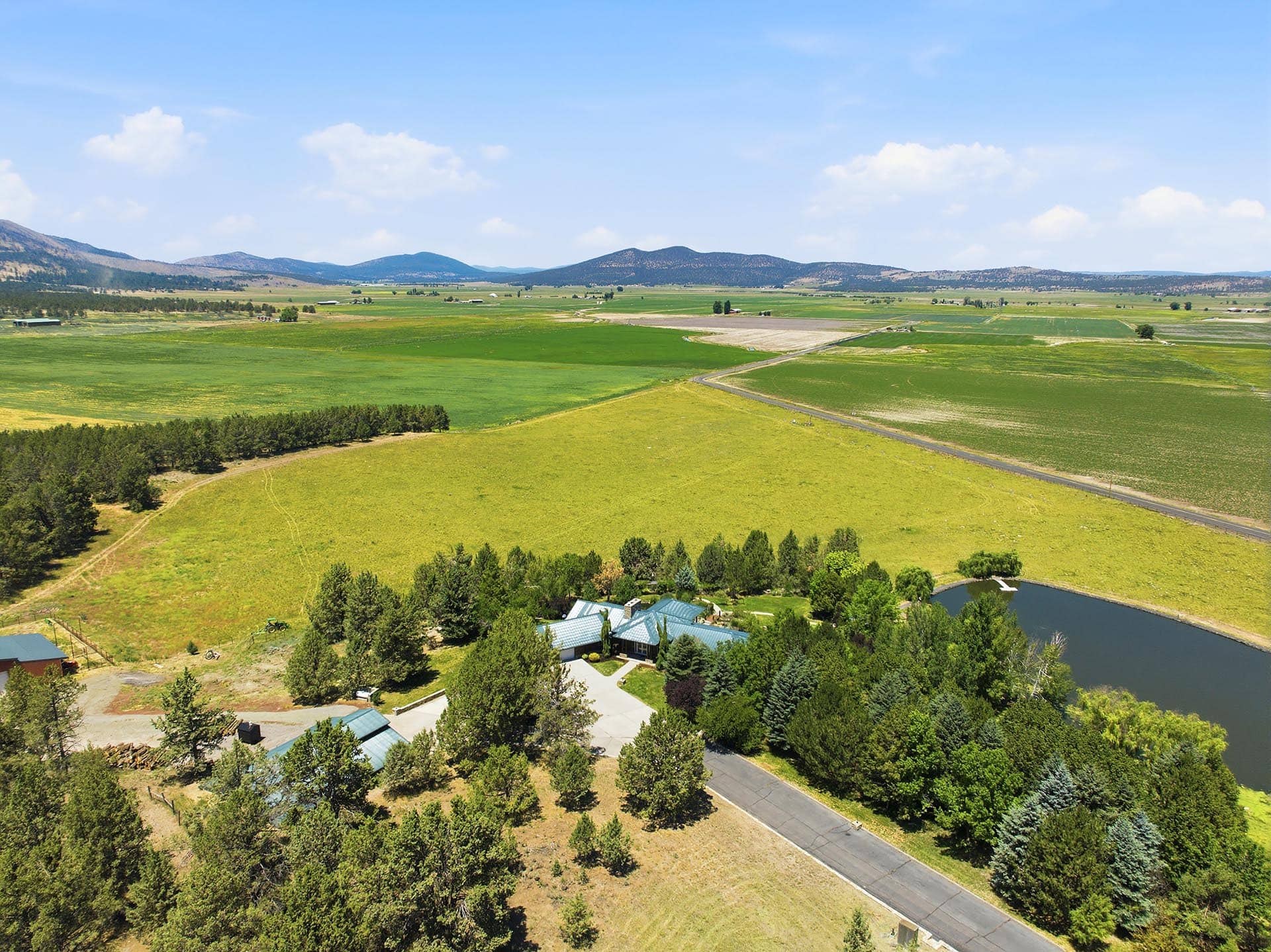 Aerial view of green fields, roads, and distant hills with a cluster of buildings by a tree-lined edge and a small pond in the foreground