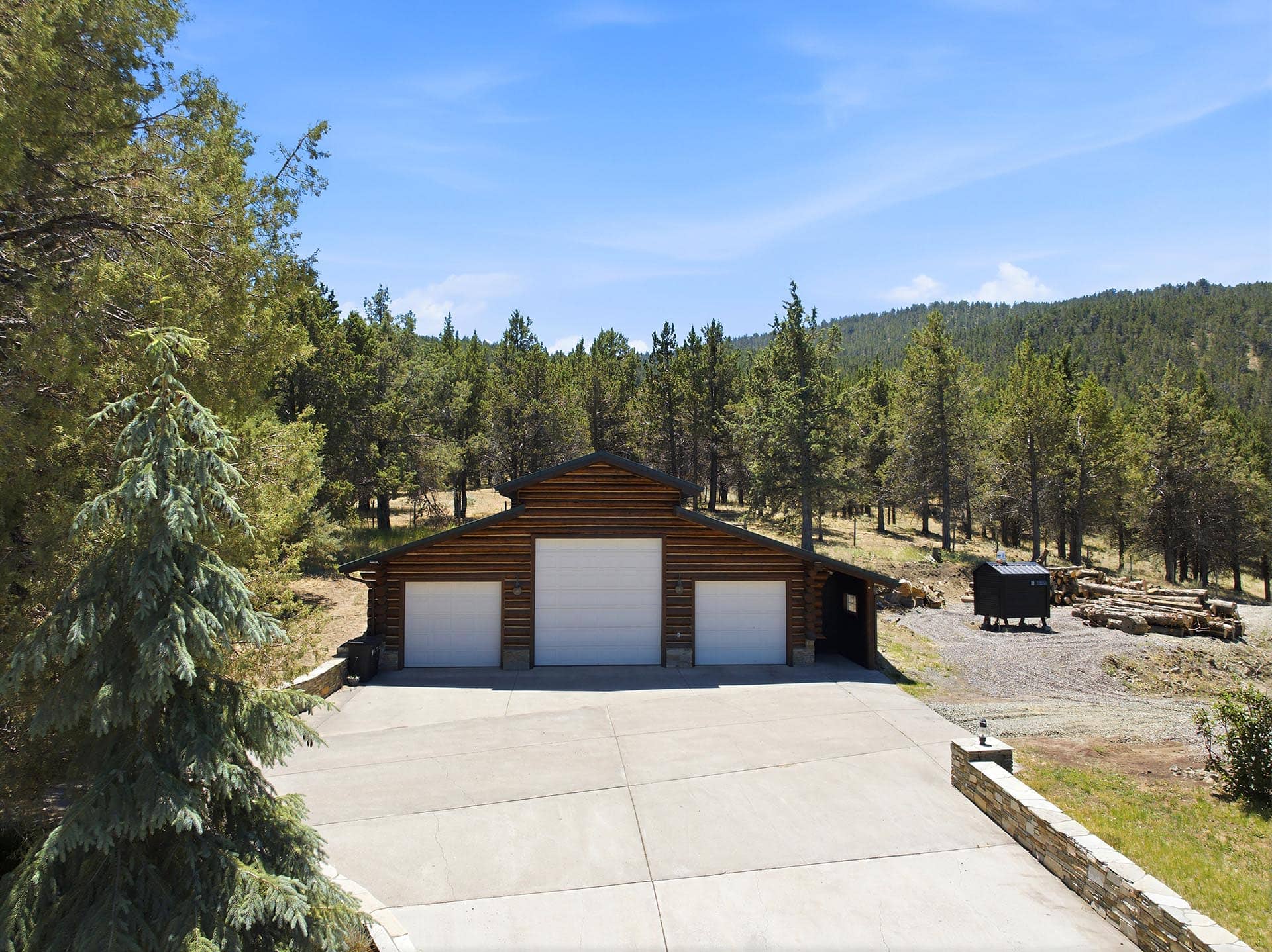 Wood cabin with three white garage doors in a concrete driveway, surrounded by trees under a clear blue sky