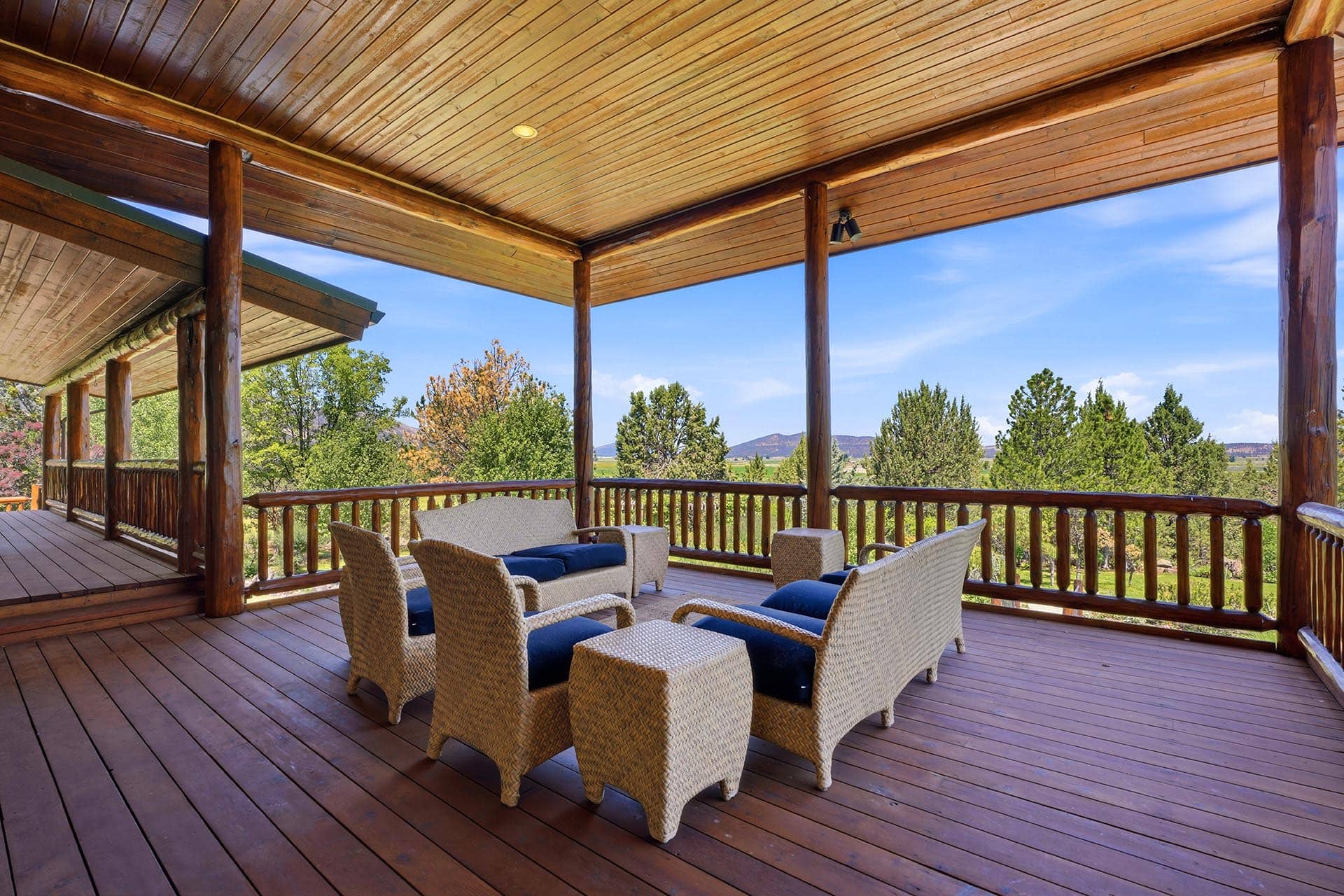 Covered wooden deck with log railings and wicker seating overlooking a forested landscape under a clear blue sky.