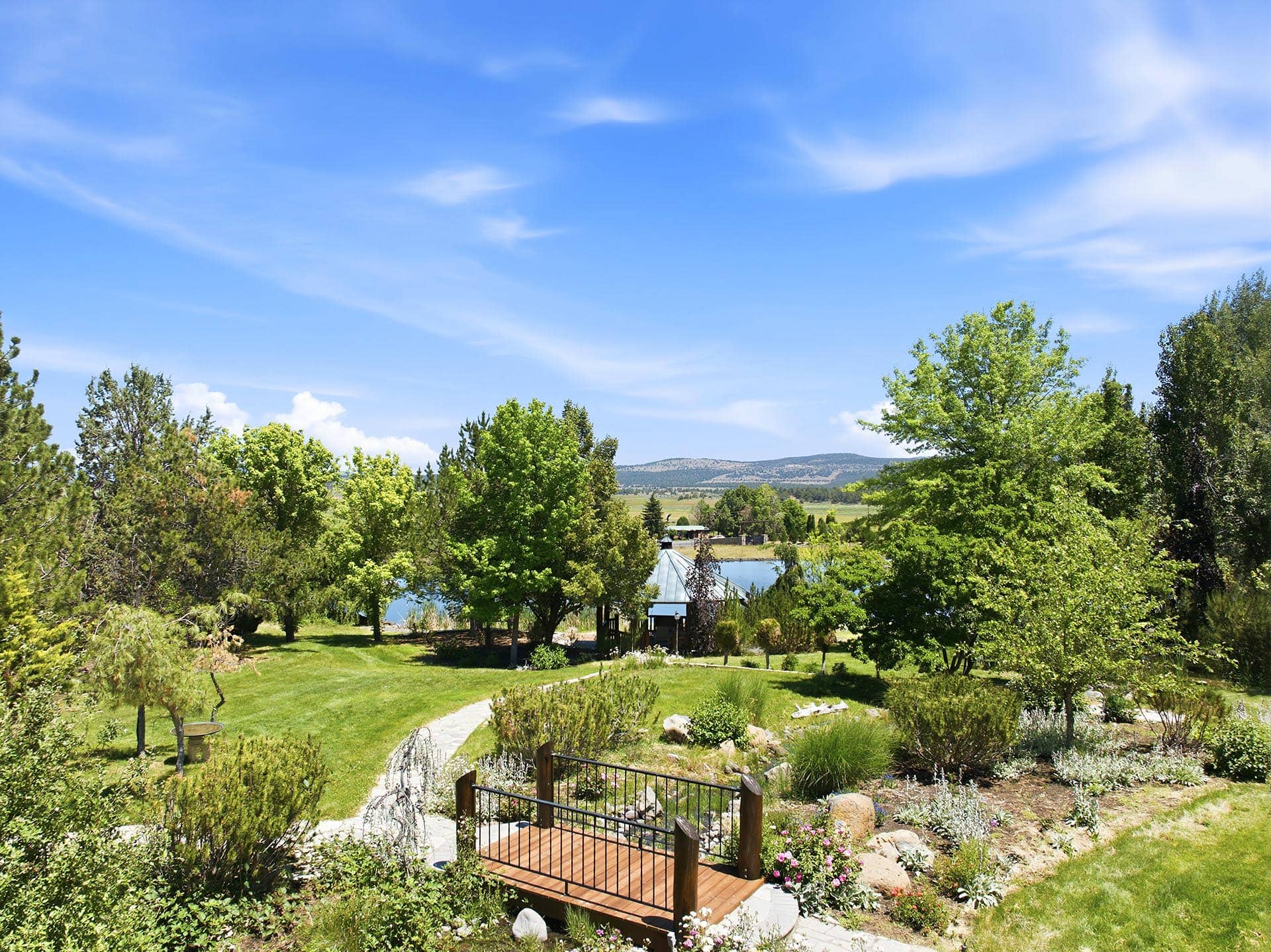 Backyard garden with a wooden deck railing in the foreground, green lawn, trees, and distant hills under a blue sky with wispy clouds.