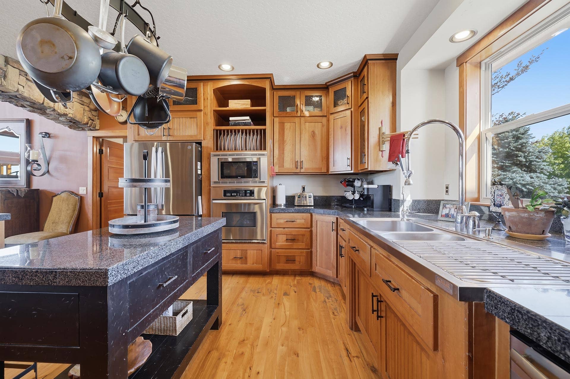 Bright kitchen with wooden cabinets, granite countertops, and a hanging pot rack above an island with dark drawers.
