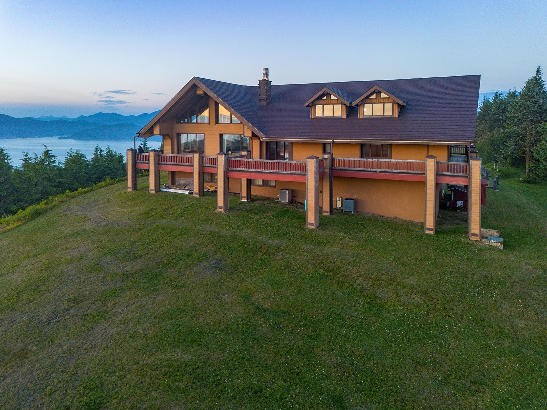 Two-story wooden house on a grassy hillside with a wraparound balcony, overlooking a forest, lake and distant mountains.