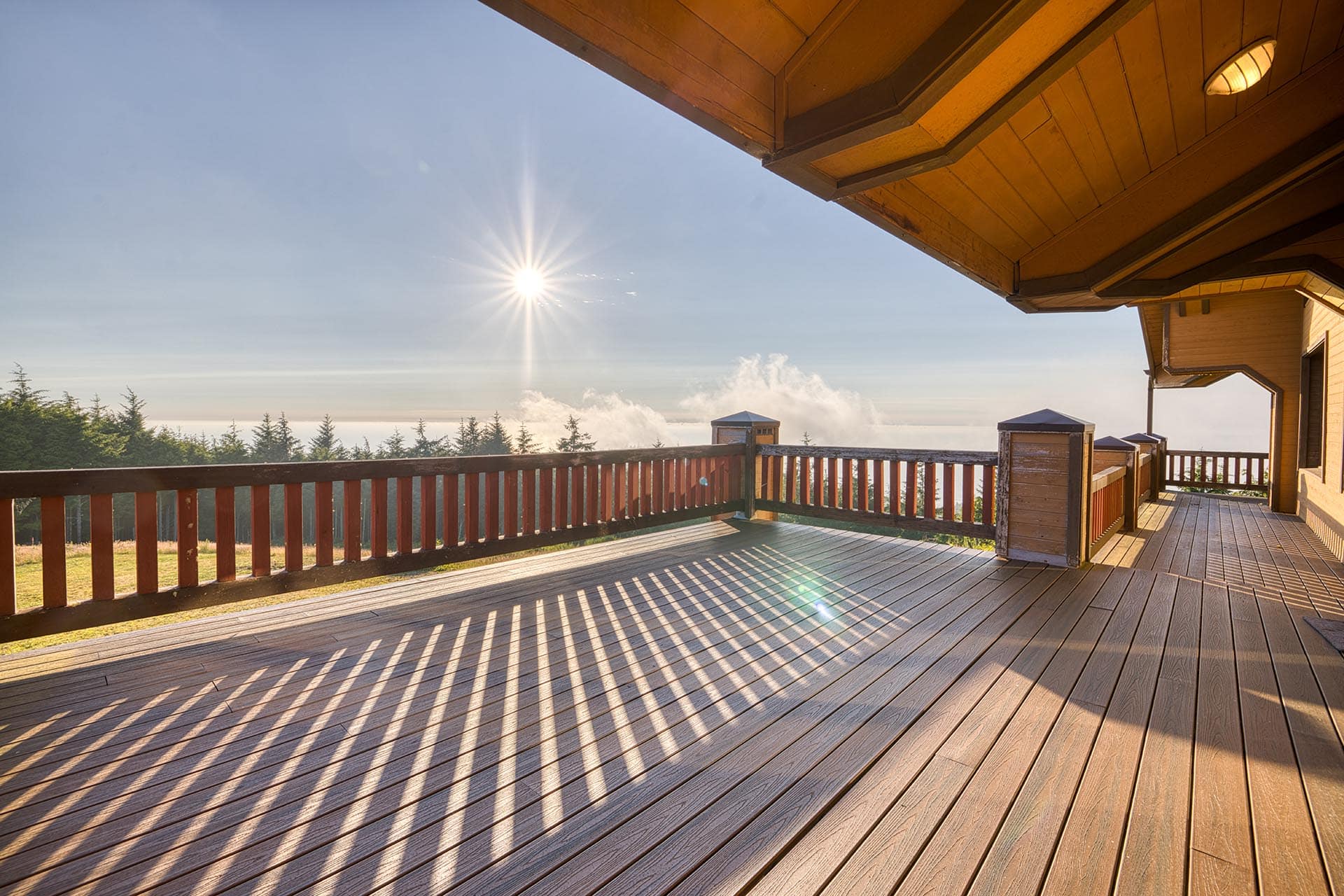 Sunlit wooden deck with a railing, long shadows cast by the railing across the planks and a distant treeline.
