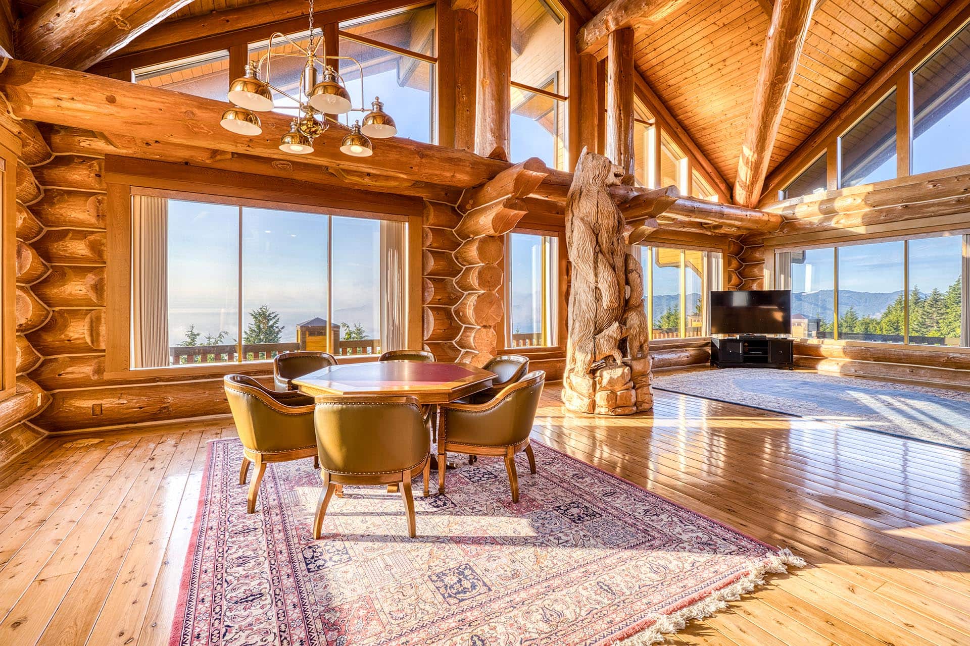 Sunlit log cabin interior with large windows, exposed wooden beams, and a round dining table surrounded by four chairs on a patterned rug.