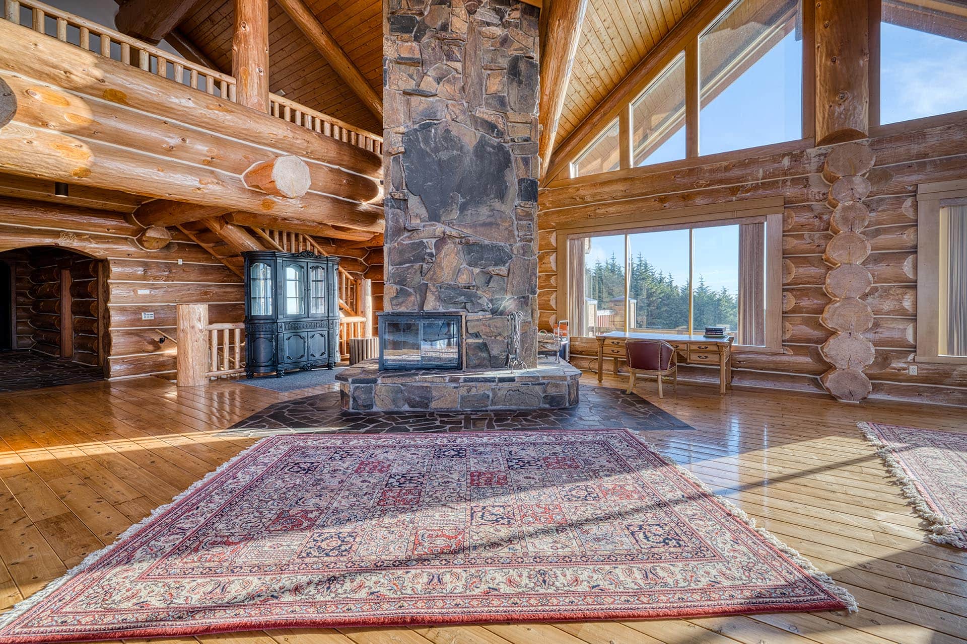 Rustic log cabin living room with a tall stone fireplace, exposed wooden beams, and large windows letting in daylight over a patterned area rug.