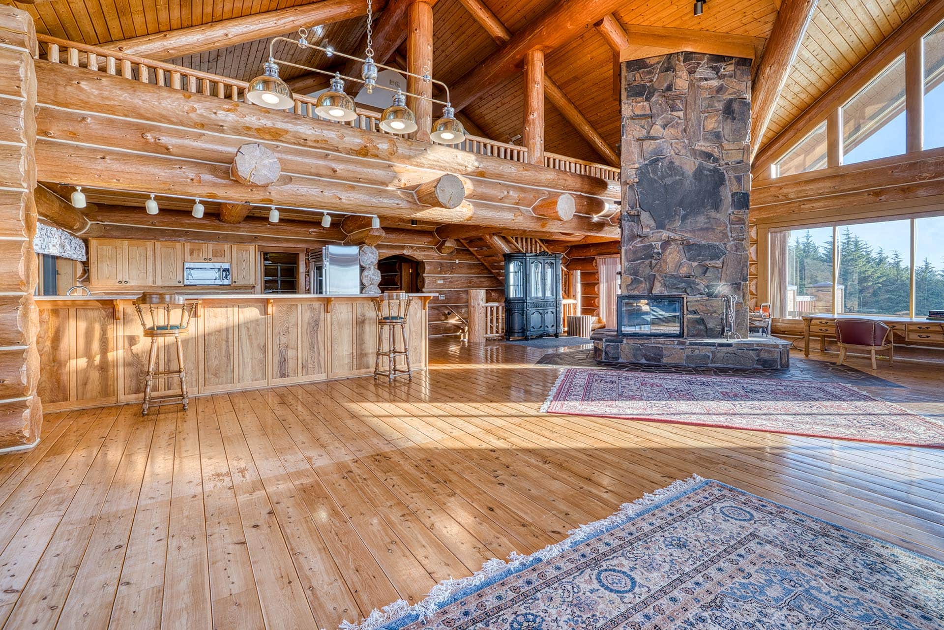 Spacious log cabin interior with a kitchen bar, exposed timber beams, and a large stone fireplace under a vaulted ceiling.
