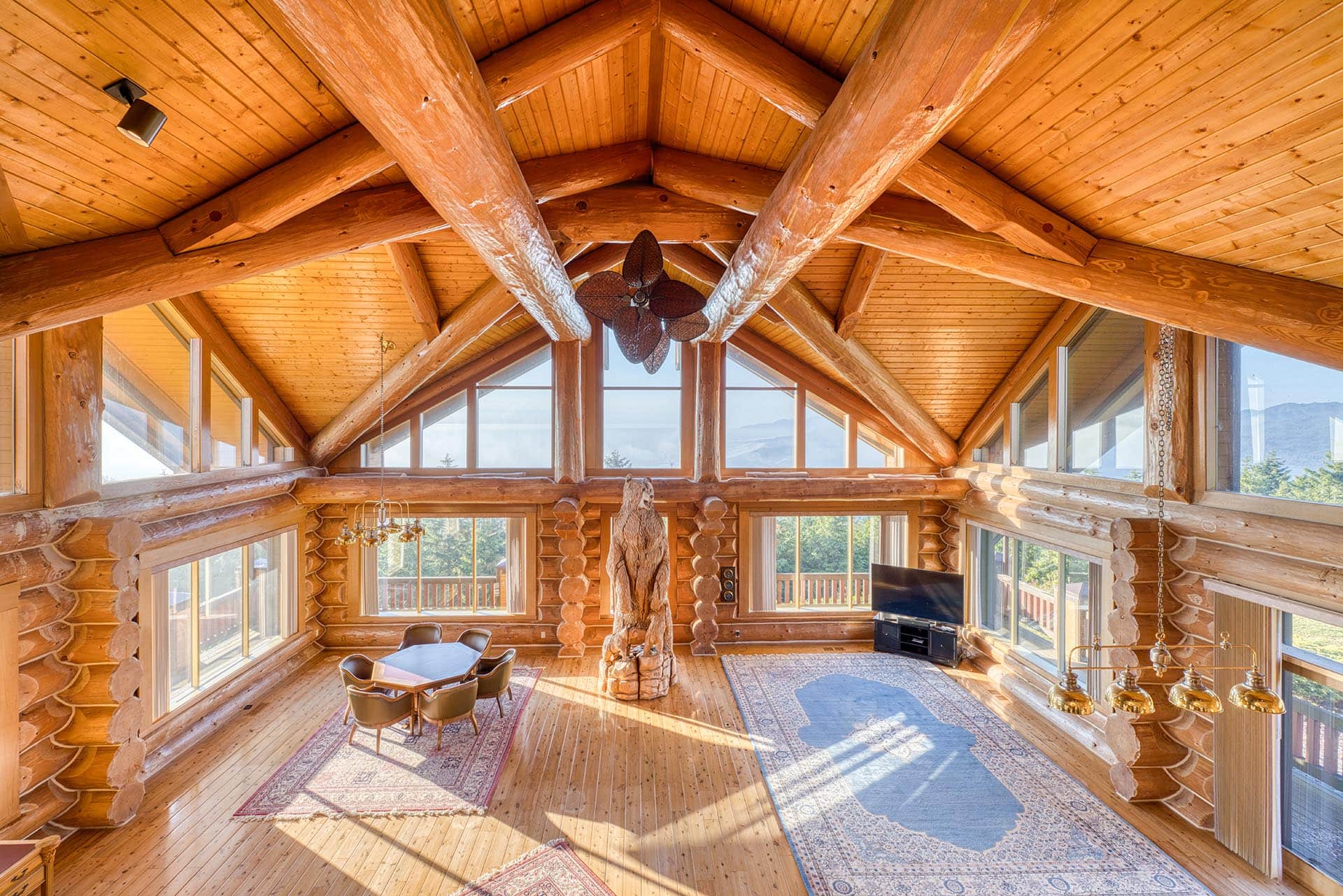 Rustic log cabin living room with a vaulted wooden ceiling and exposed log beams, large windows, and a seating area.