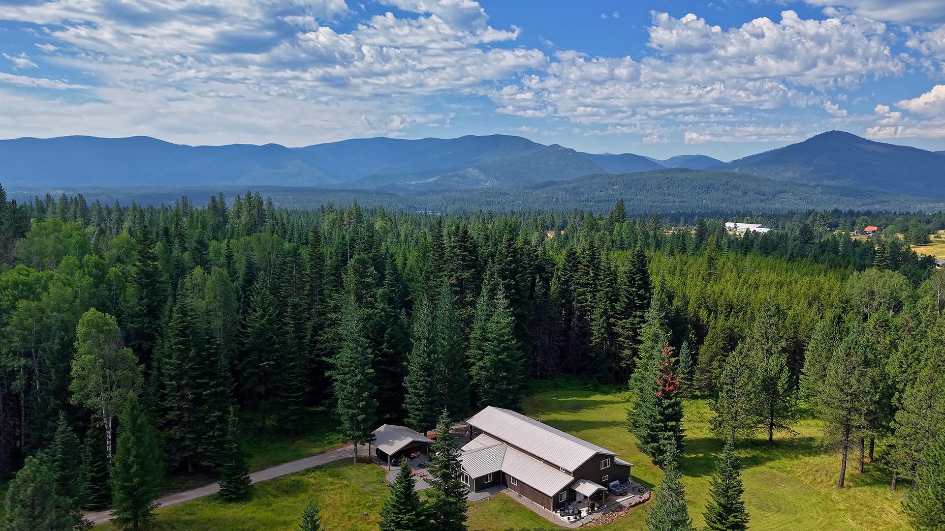 Valley framed by dense evergreen forest with a few buildings, and blue mountains on the horizon under a cloudy sky.