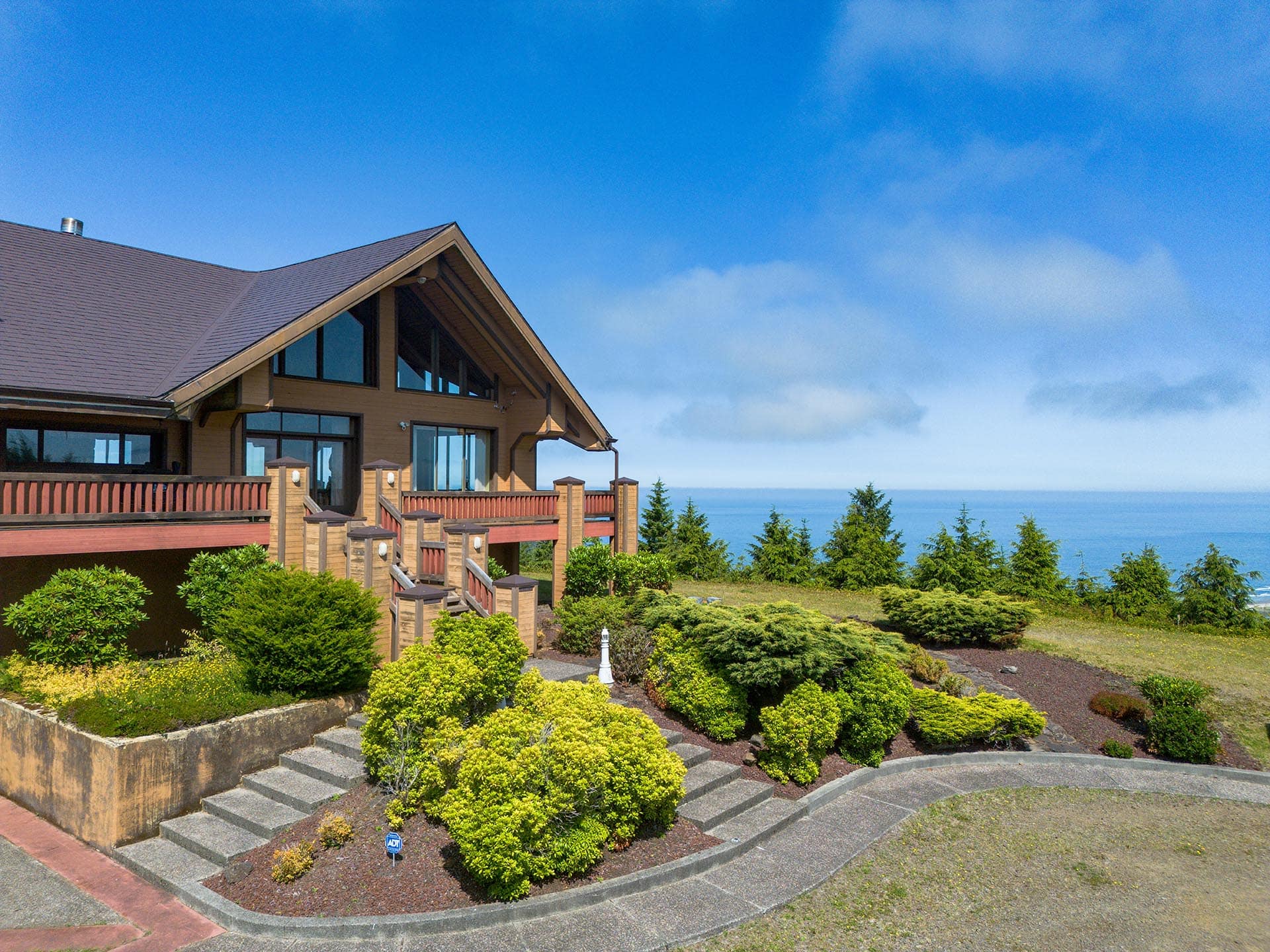 Modern wooden house with a steep gabled roof overlooking the ocean, surrounded by manicured shrubs and a curved stone path.