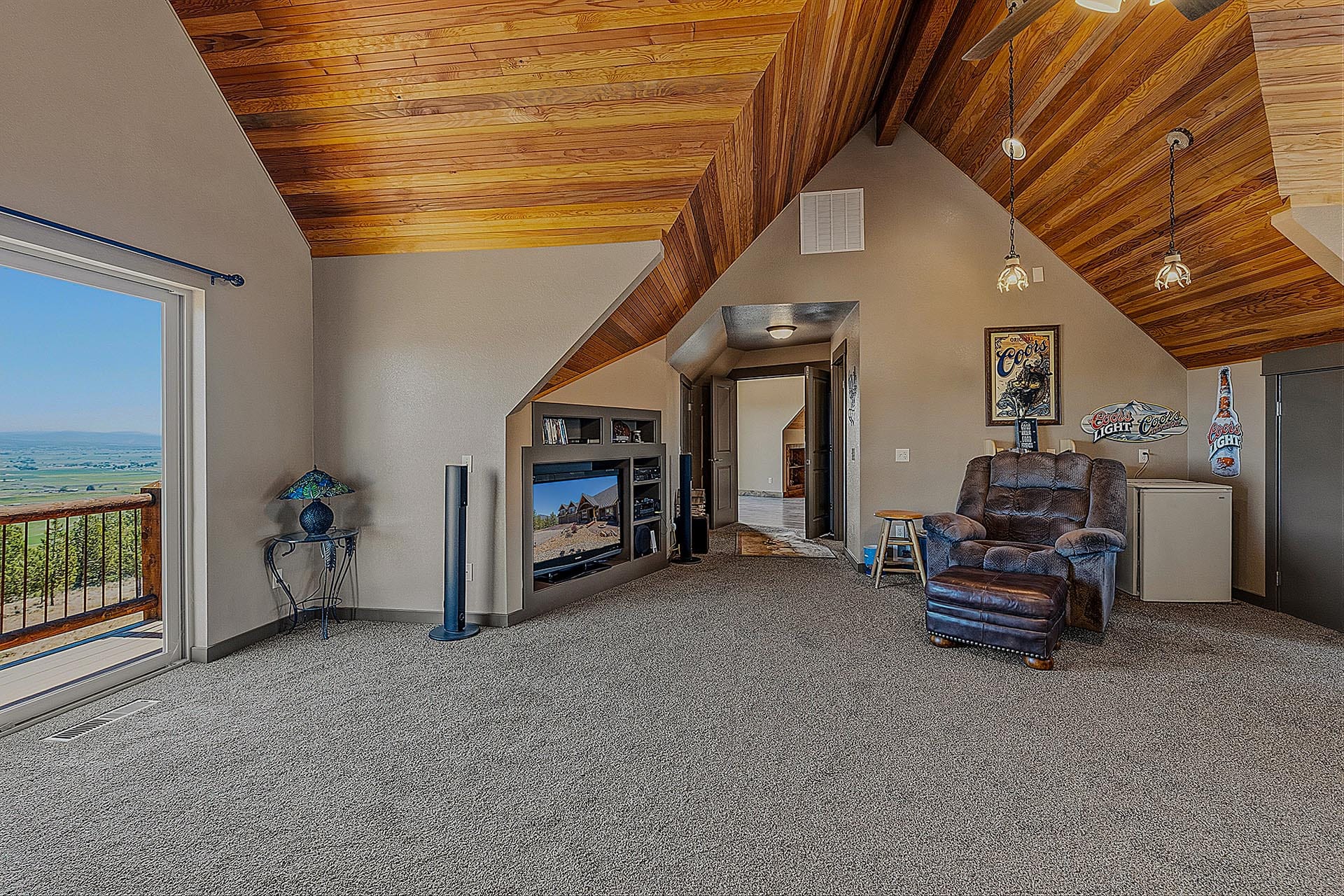 Loft-style living room with a vaulting wooden ceiling, tall glass door to a balcony, and a distant rural landscape view behind a railing.