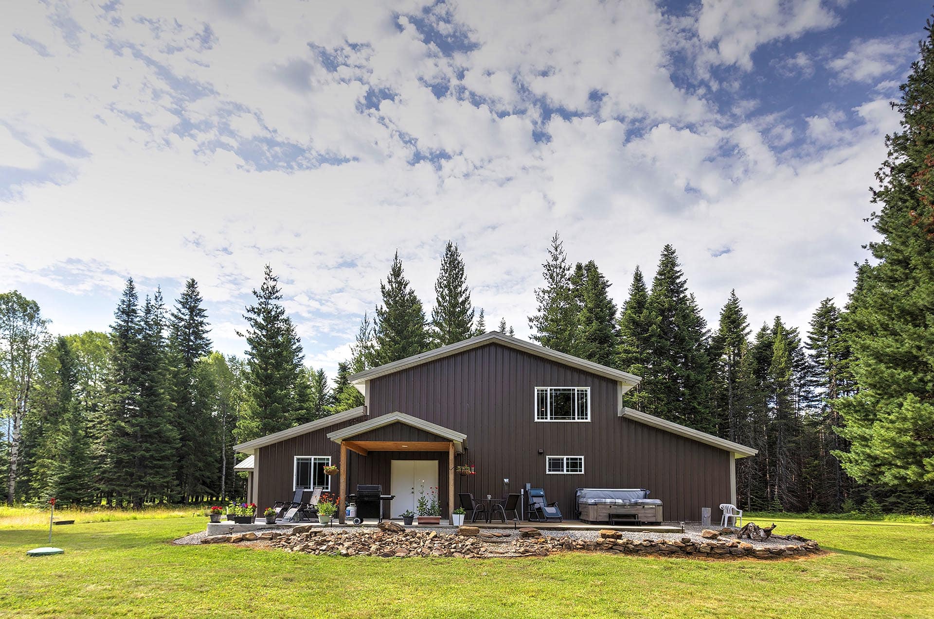 Brown two-story house with a front porch, patio seating, grill, and potted plants in a grassy yard surrounded by tall pine trees under a blue sky with clouds.