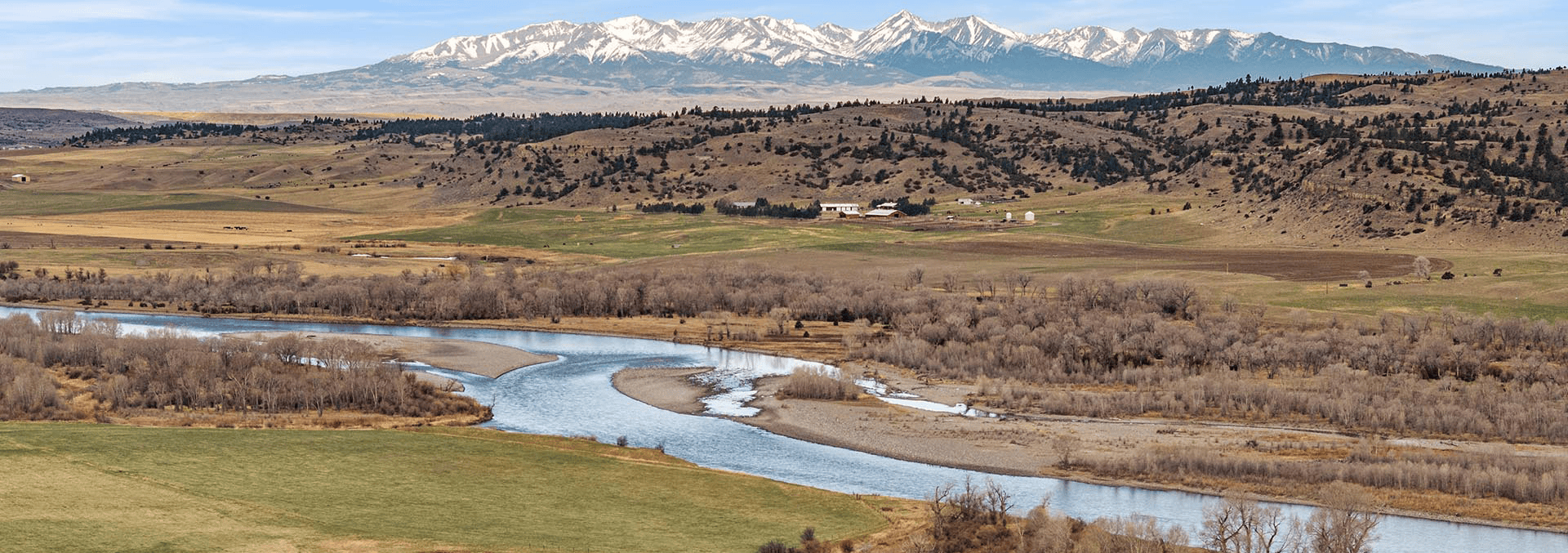 Snow-capped mountains rise in the distance over rolling farmland, with a winding river and leafless trees in the foreground.