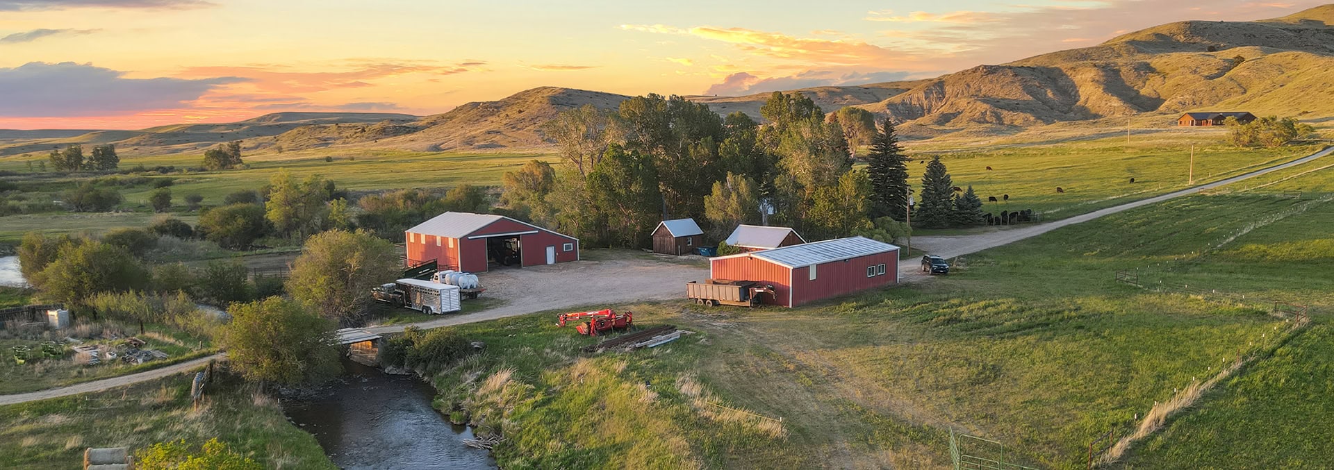 Sunset over a rural farm with red barns, green fields, and distant hills beside a winding river at dusk.