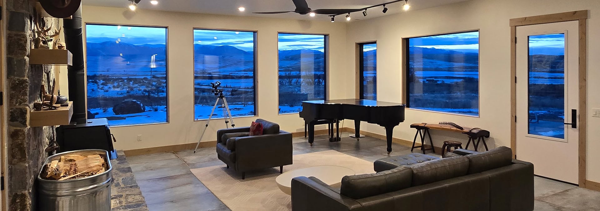 Cozy music room with a grand piano, two gray sofas, and a telescope by large windows showing snowy mountains outside.