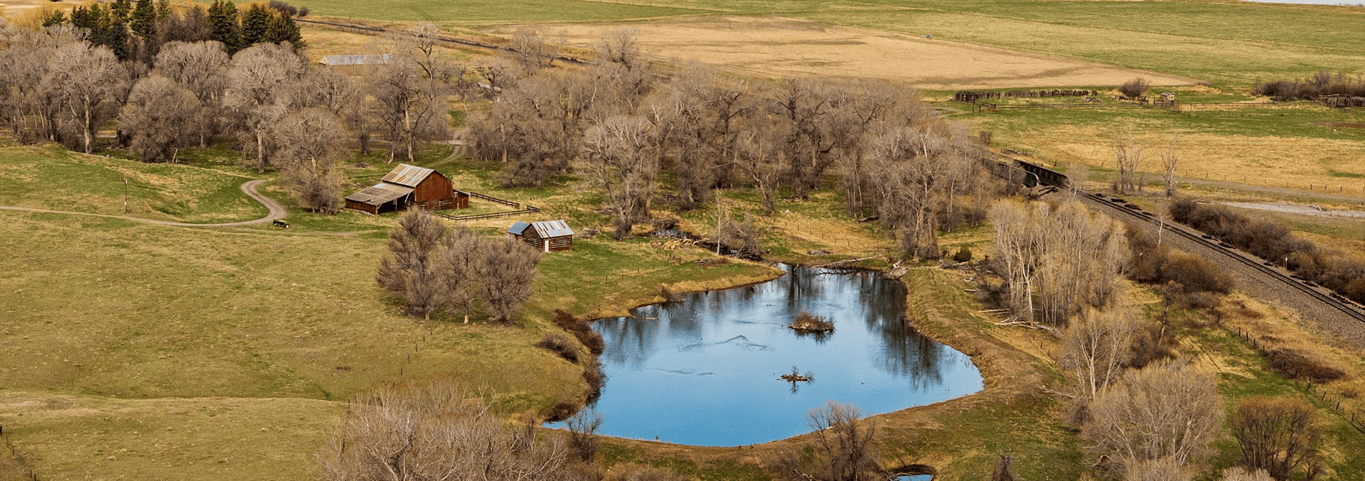 A rural farmstead with a pond, leafless trees, red barns, and a railroad track along the fields (aerial view).